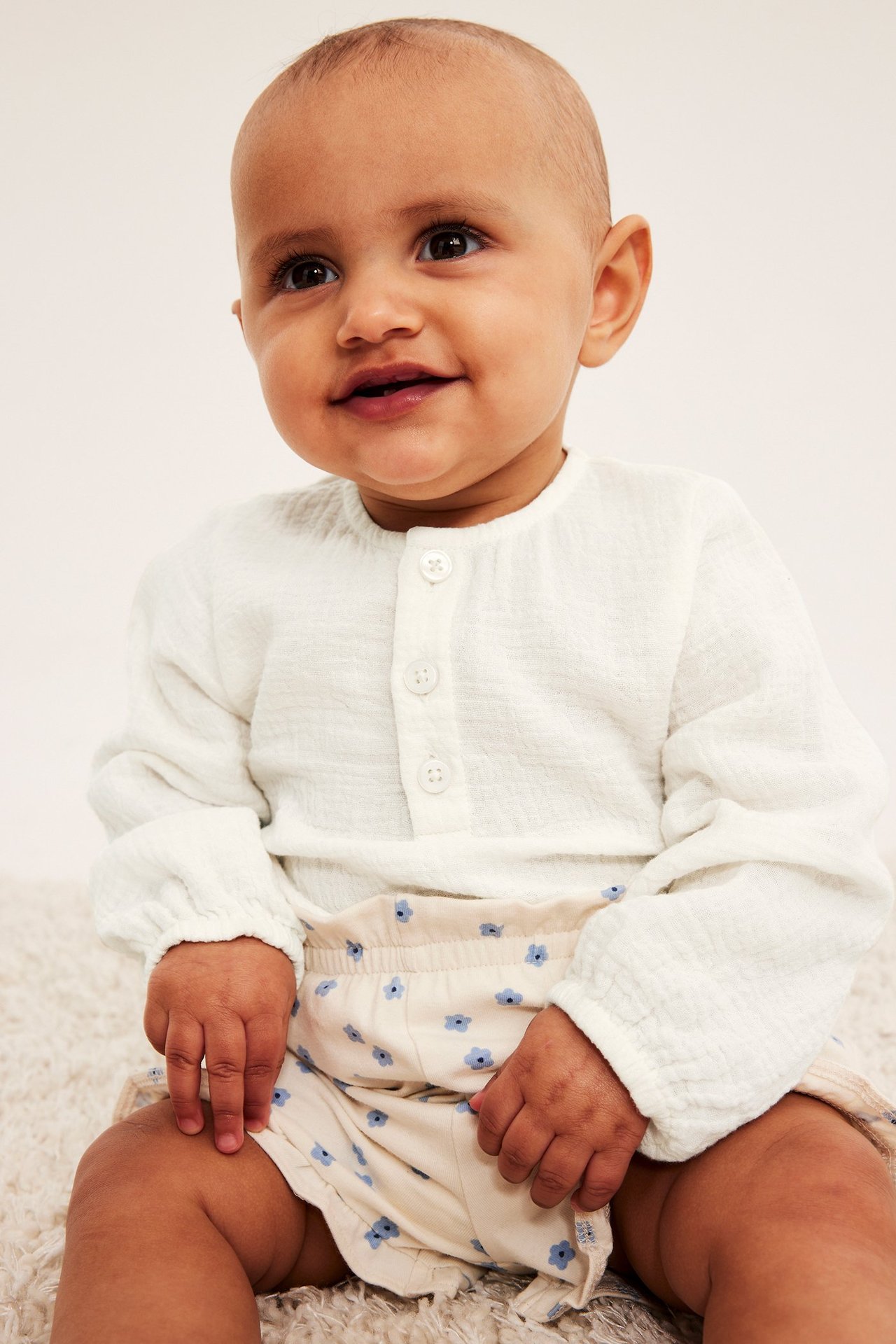 Framifrån, en baby bär en offwhite långärmad muslinbody med knappar och beige blommiga shorts.