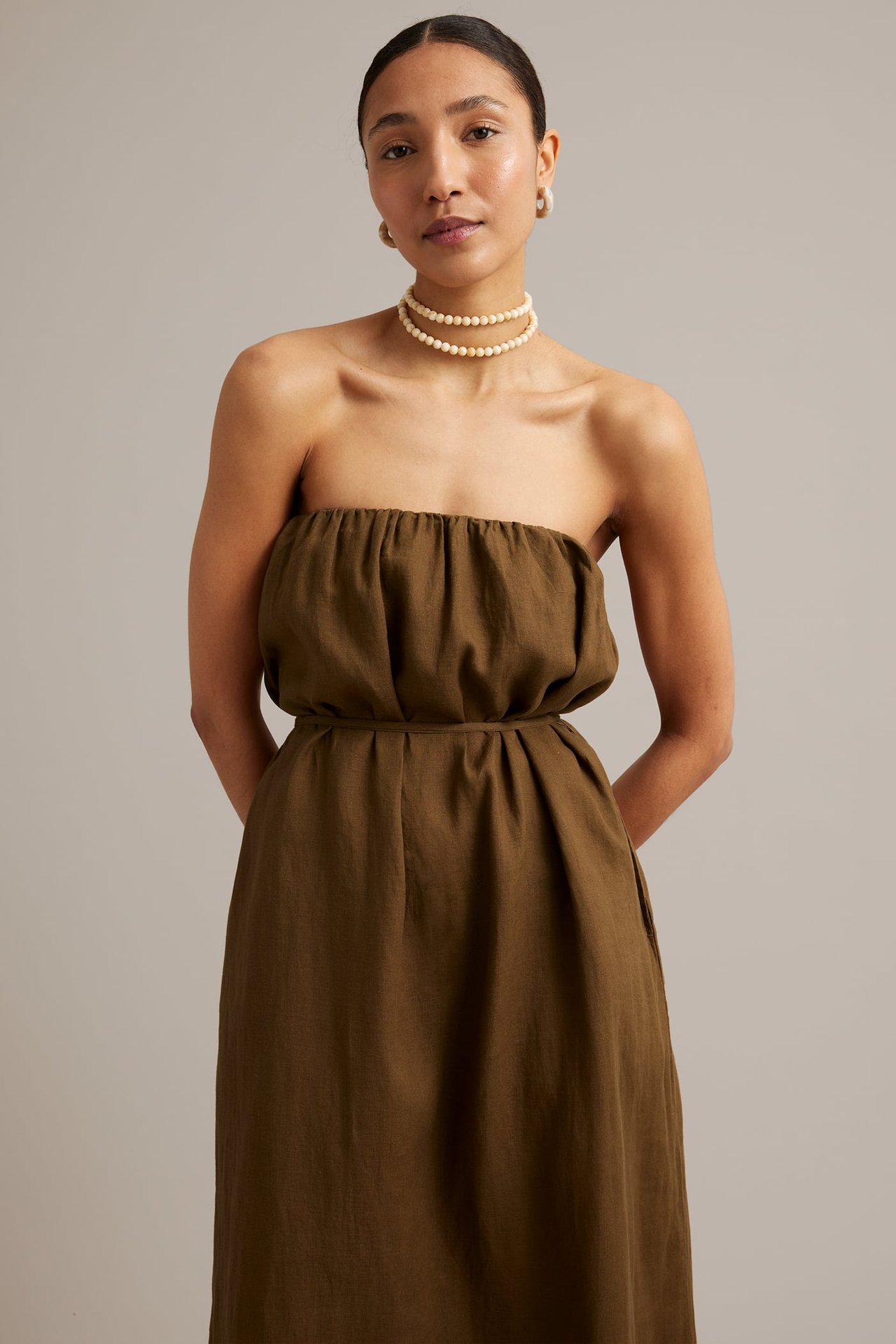 Front view: Kvinna i brun linne strapless klänning med knytskärp, pärlhalsband och krämfärgade örhängen.