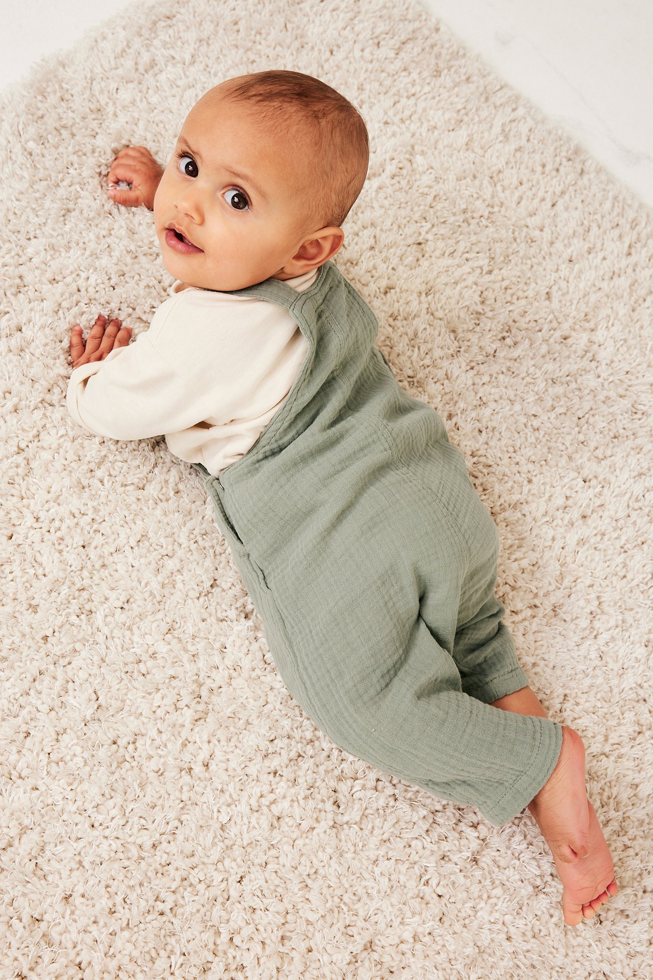 Ylhäältä kuvattu vauva yllään yksiväriset tummanvihreät musliinihaalarit ja luonnonvalkoinen pitkähihainen paita.