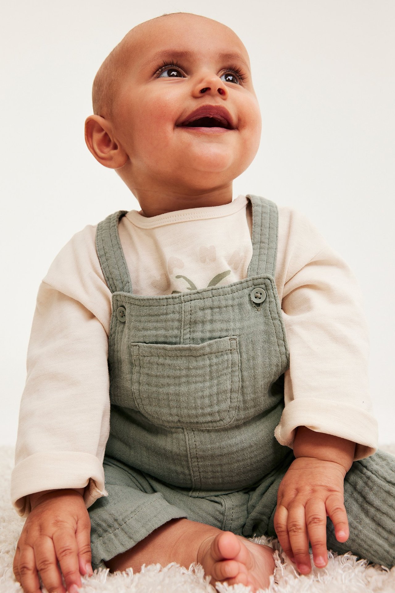 Forfra, smilende baby i mørkegrønne muslinselebukser med knapper over en lys beige langermet t-skjorte.