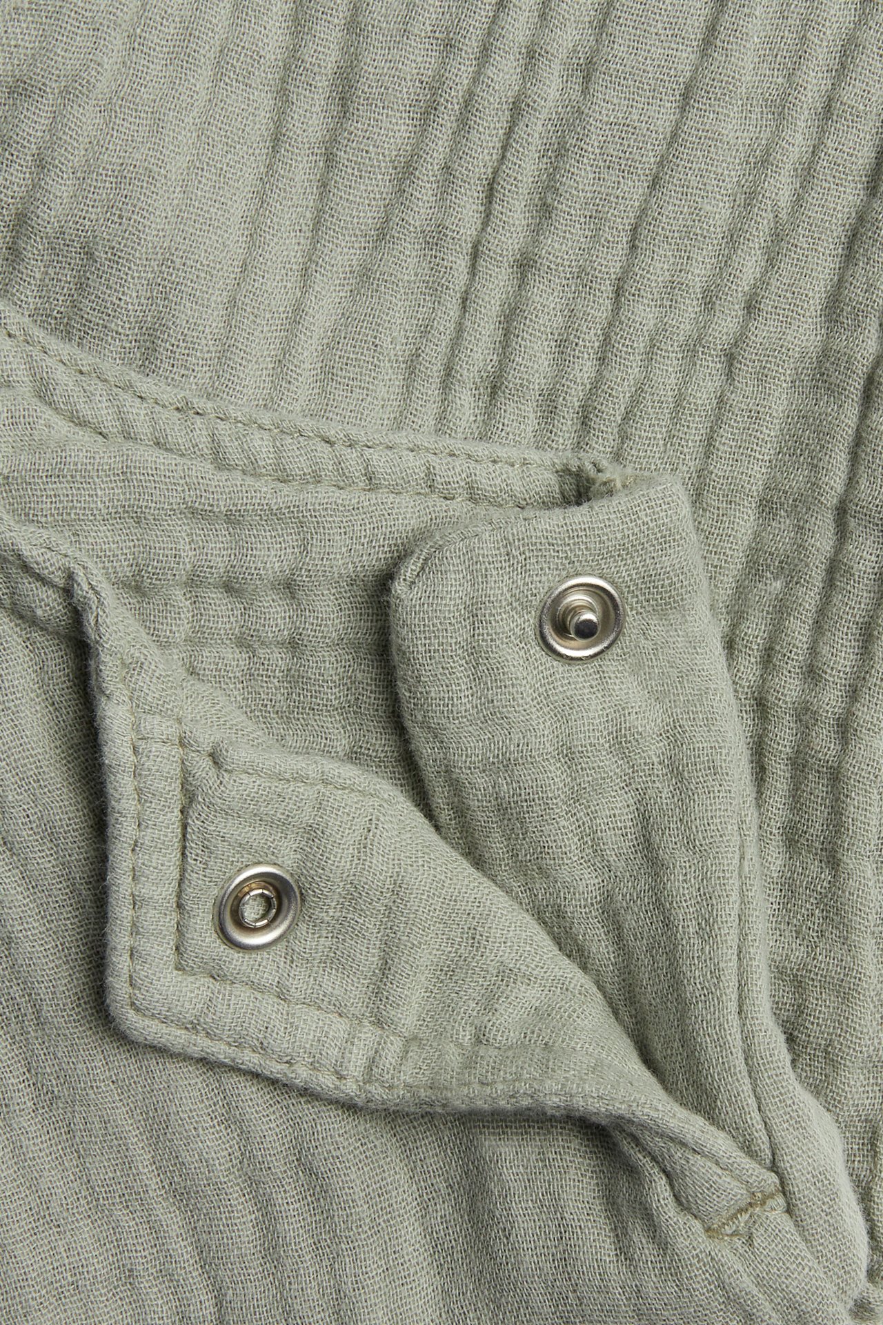 Lähikuva tummanvihreistä musliinihaalarista vauvalle, jossa on kaksi hopeanväristä painonappia.