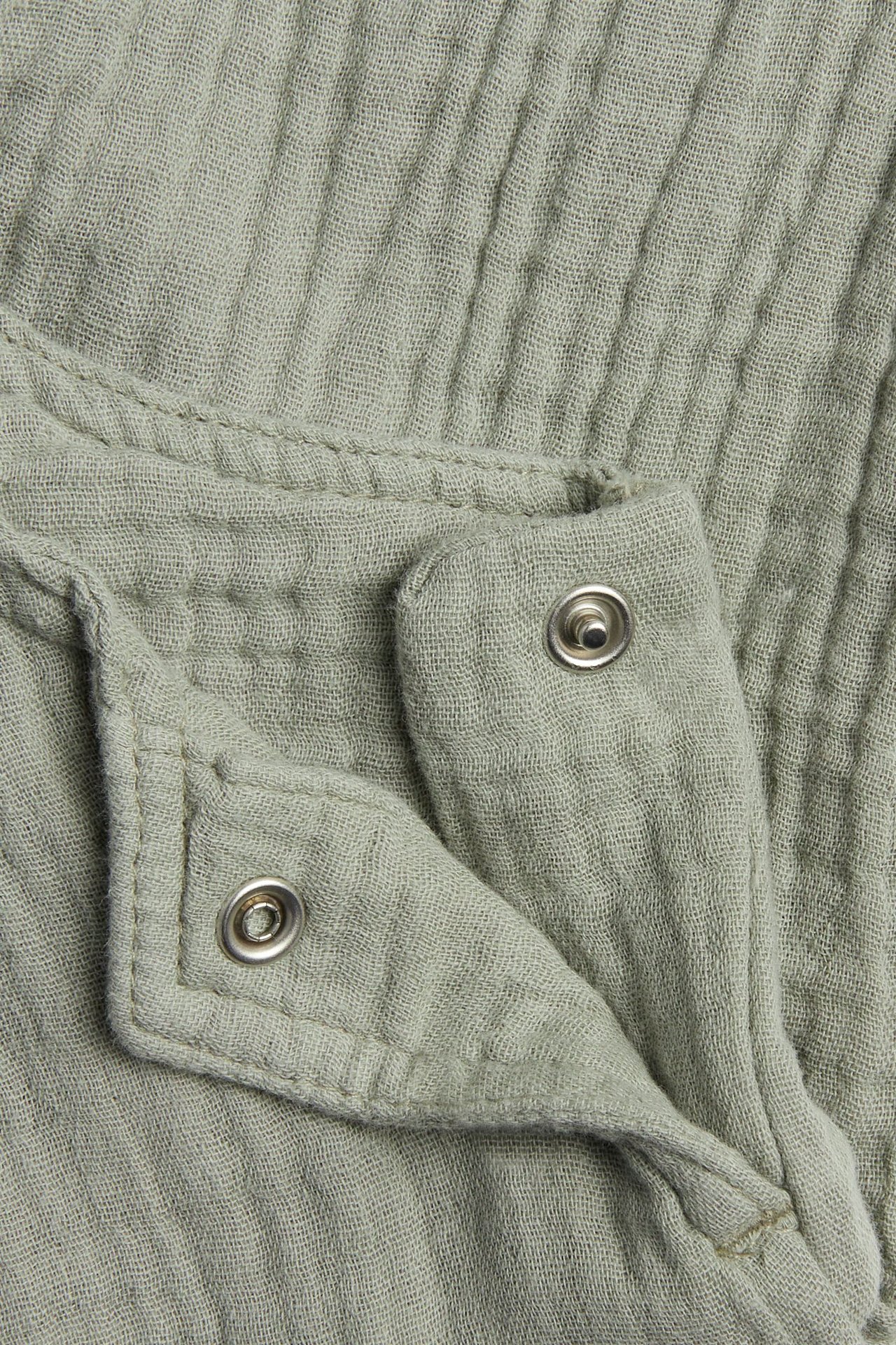 Zbliżenie na ciemnozielone muślinowe ogrodniczki niemowlęce z dwoma srebrnymi zatrzaskami.