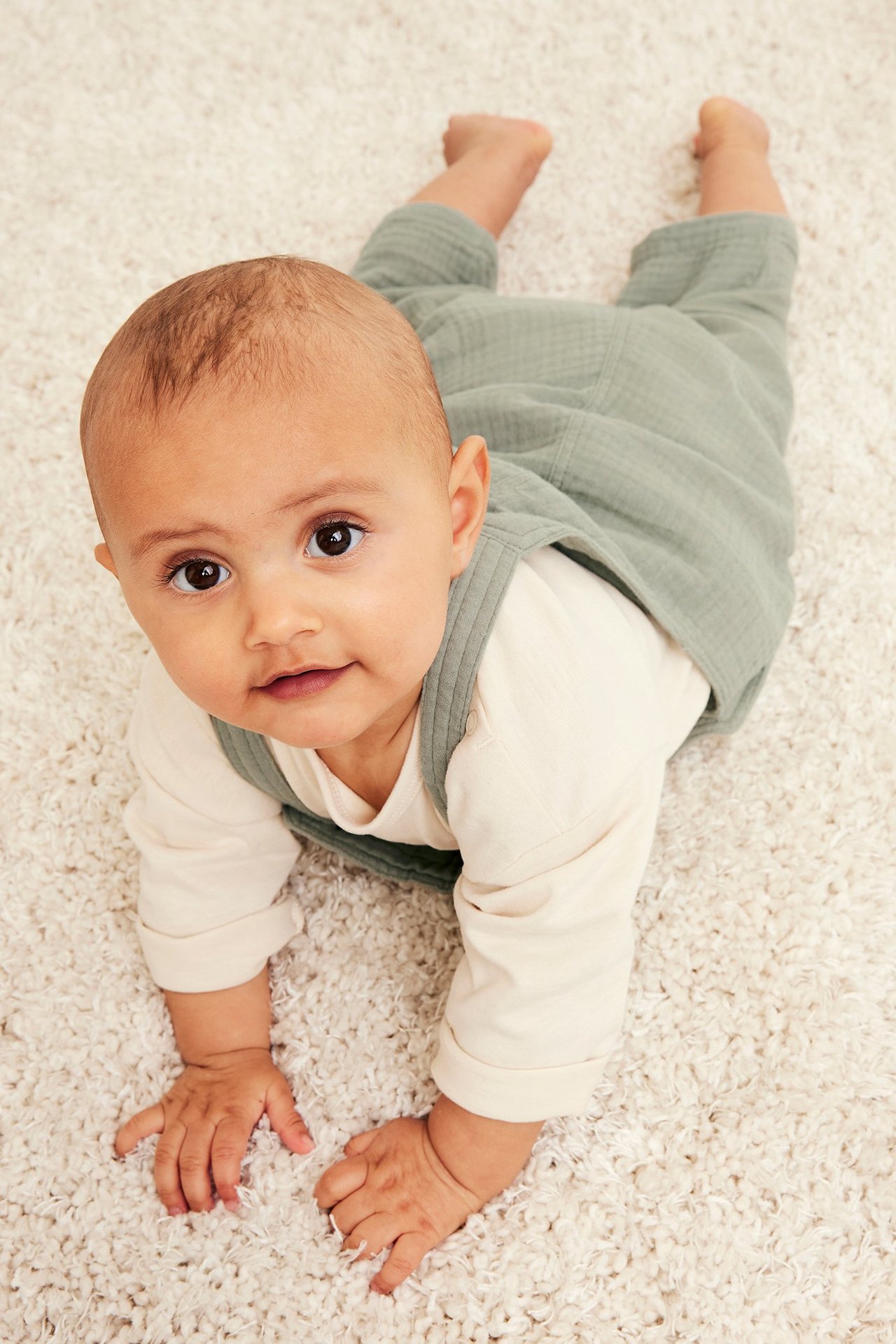 Ovenfra, forfra: en baby kryper i mørkegrønne musselin-selebukser over en lys kremfarget langermet topp.