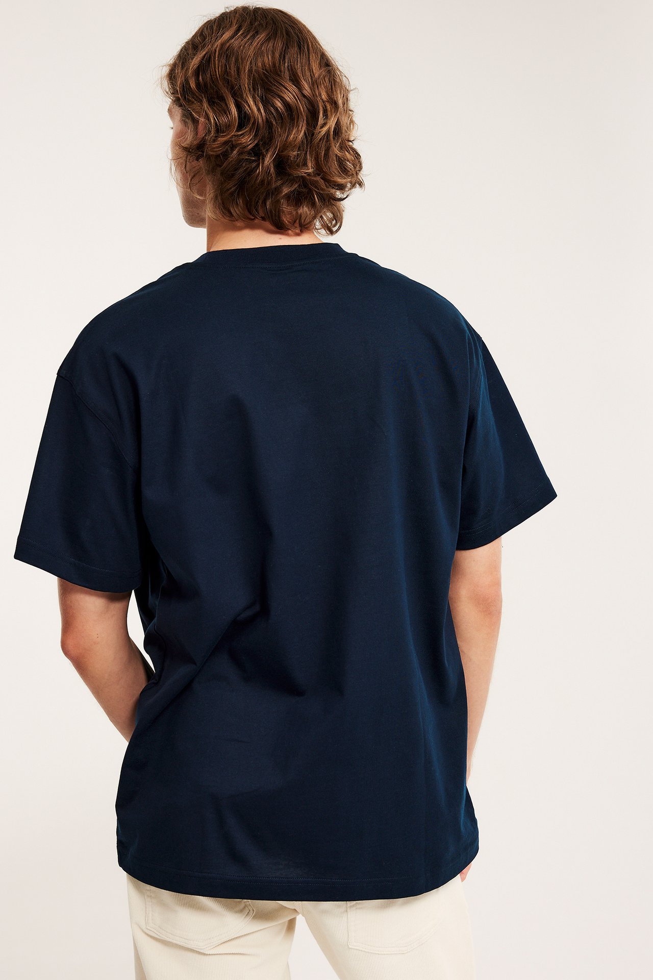 Miehen takakuva: tummansininen väljä puuvilla-T-paita ja vaaleat beiget housut.