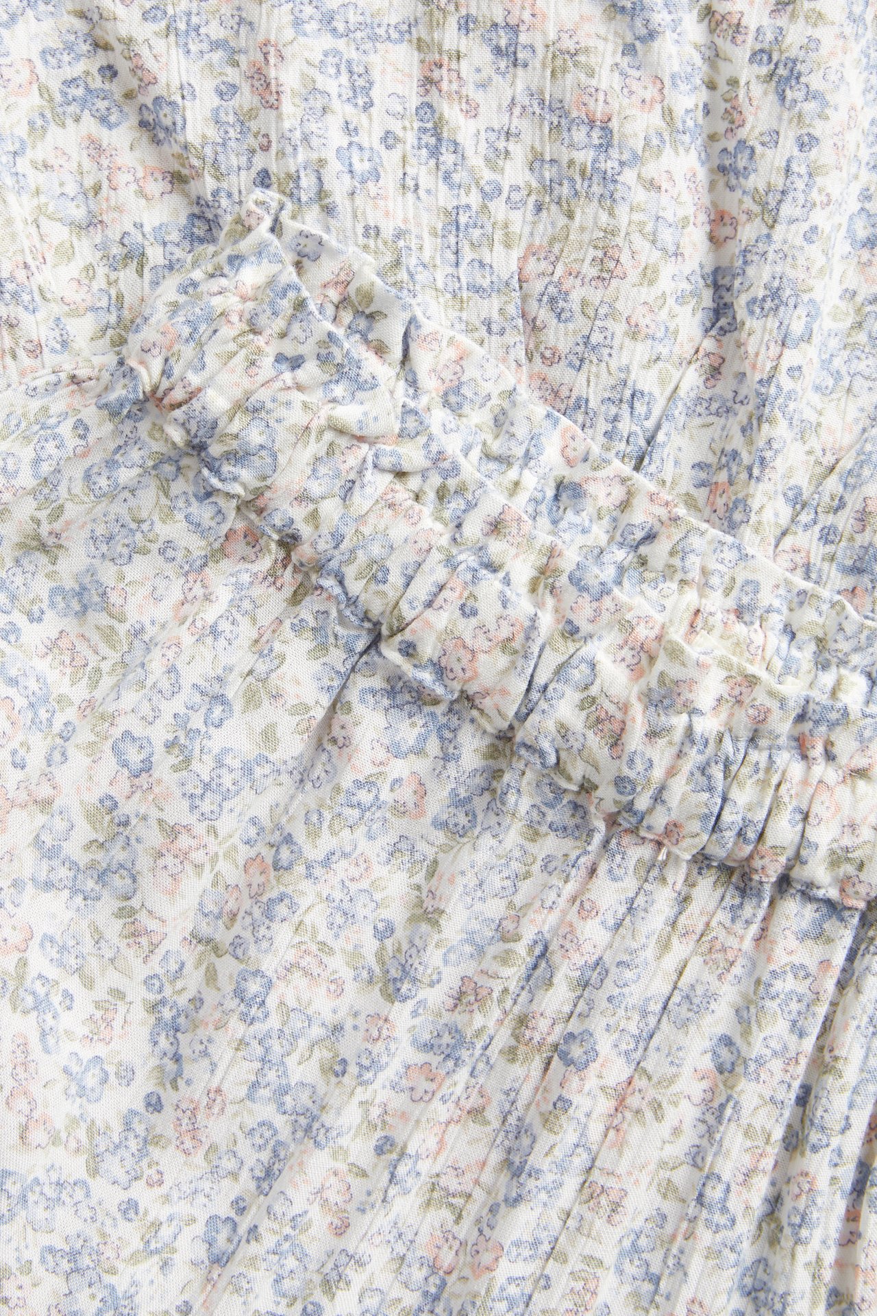 Lähikuva luonnonvalkoisesta kukkakuosisesta pitkästä hameesta, jossa pieniä sinisiä ja vaaleanpunaisia kukkia ja röyhelö.