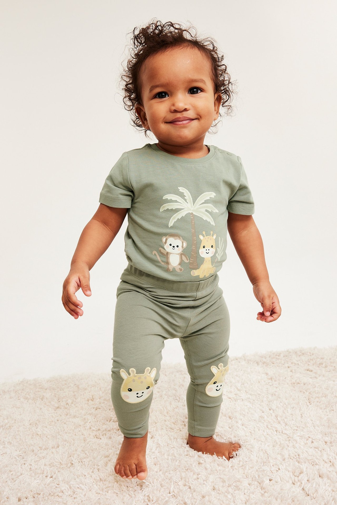 Framsida: Baby i en salviagrön kortärmad body med djurtryck och matchande leggings med giraffknäskydd.