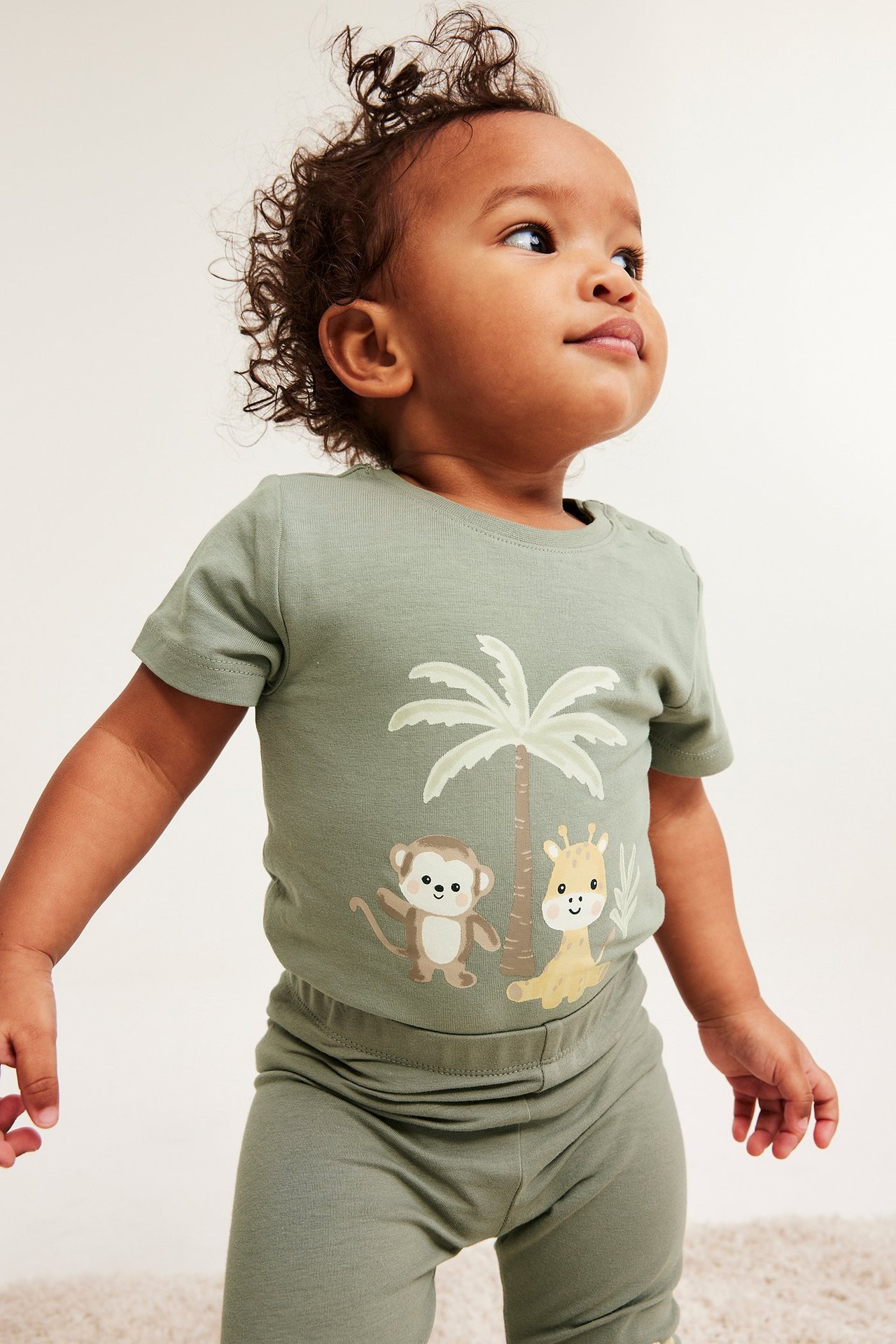 Boczny widok dziecka w ciemnozielonym body z krótkim rękawem, nadrukiem zwierzęcym i pasującymi legginsami.