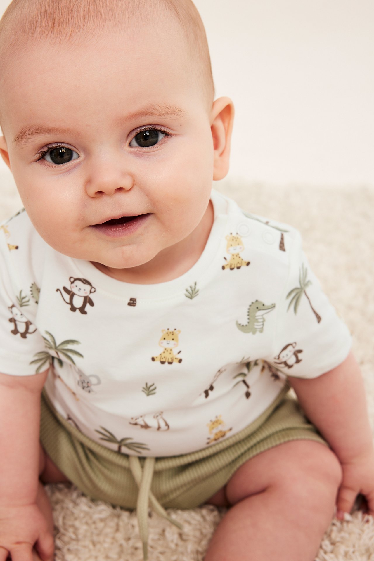 Närbild, framifrån, av en bebis i offwhite kortärmad body med djurmönster och gröna ribbade shorts.