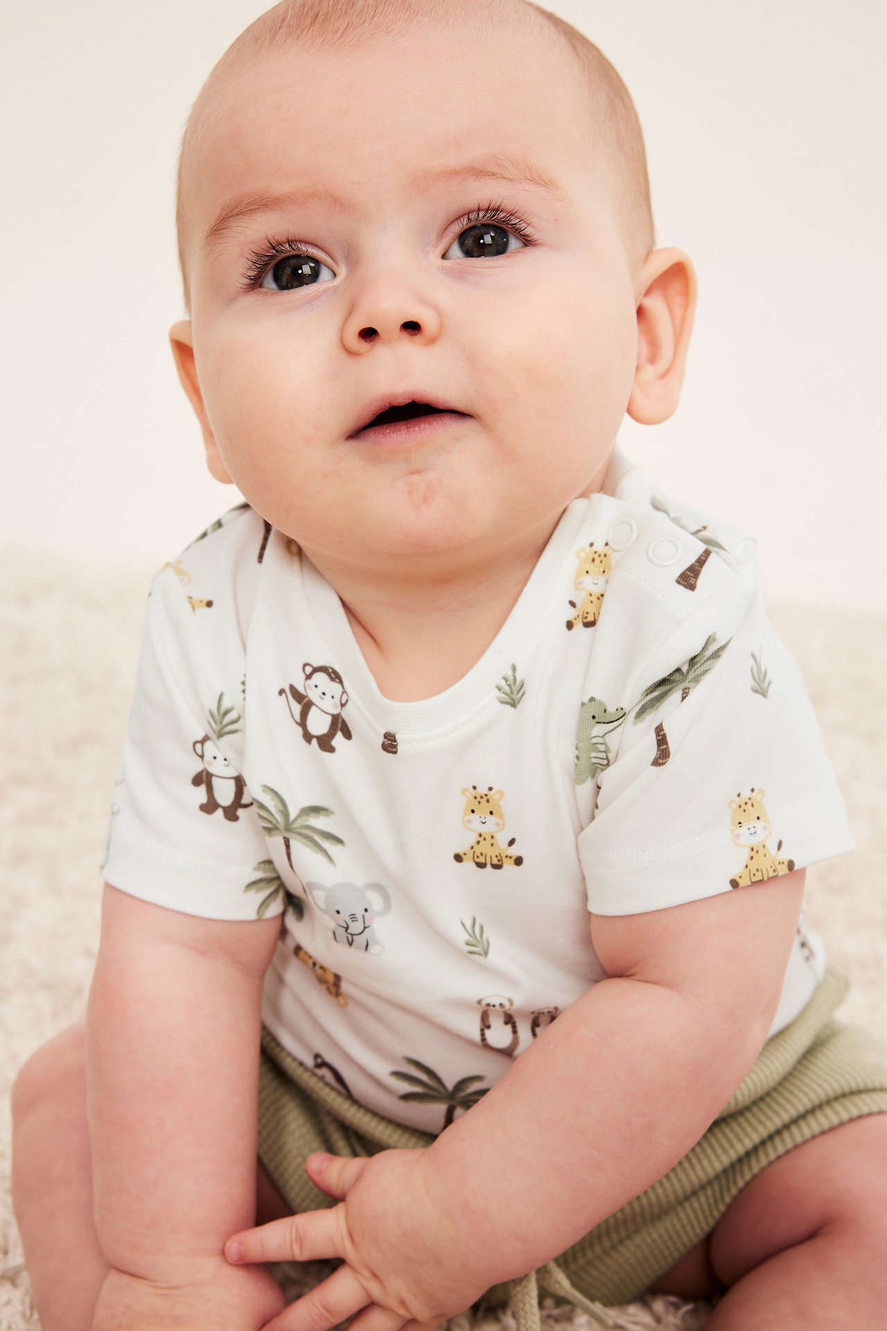 Framsida av en baby i vit kortärmad body med djurmönster, tryckknappar på axeln och gröna shorts.