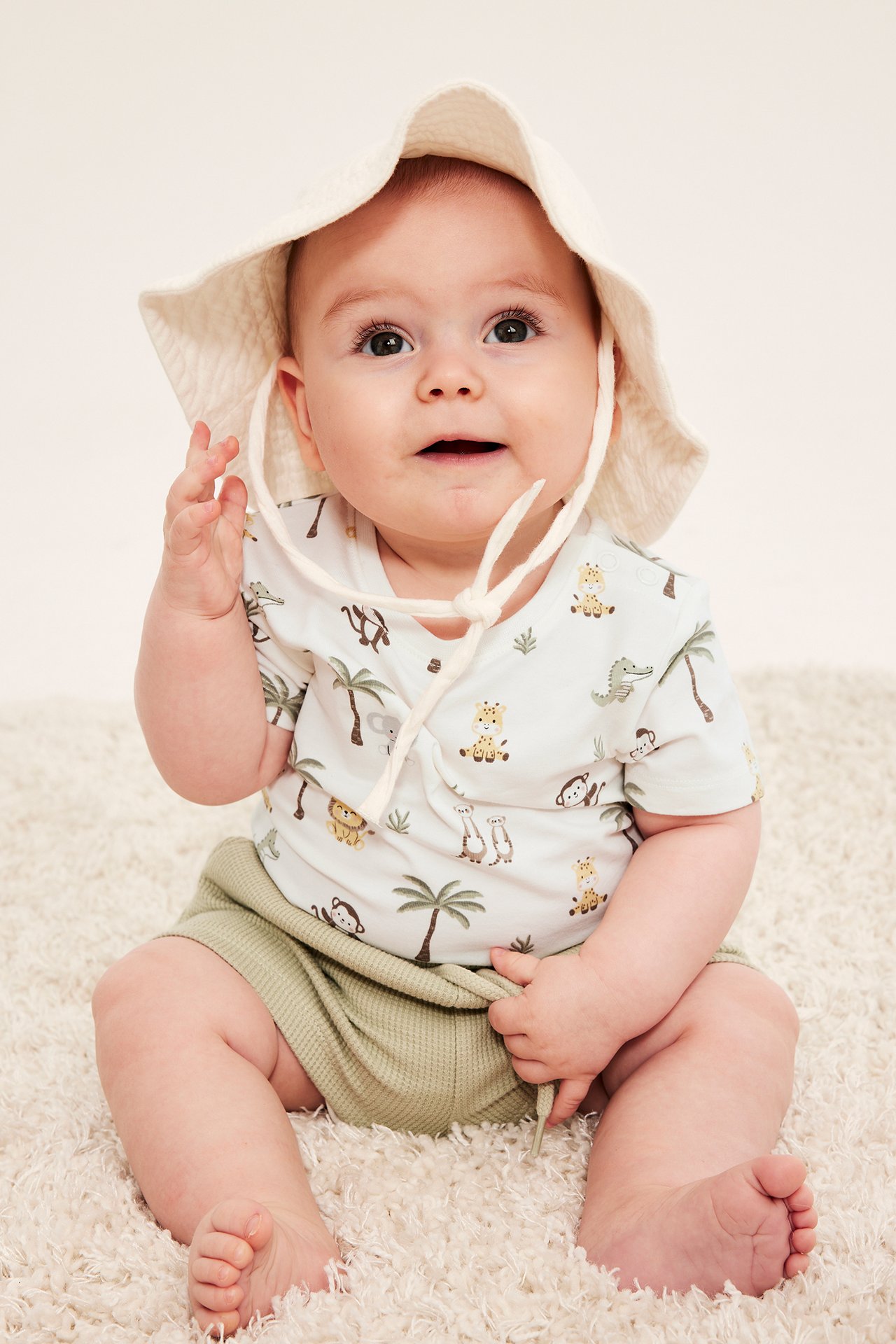 Framifrån: baby i en benvit kortärmad body med djurmönster, ljusgröna shorts och en benvit solhatt.