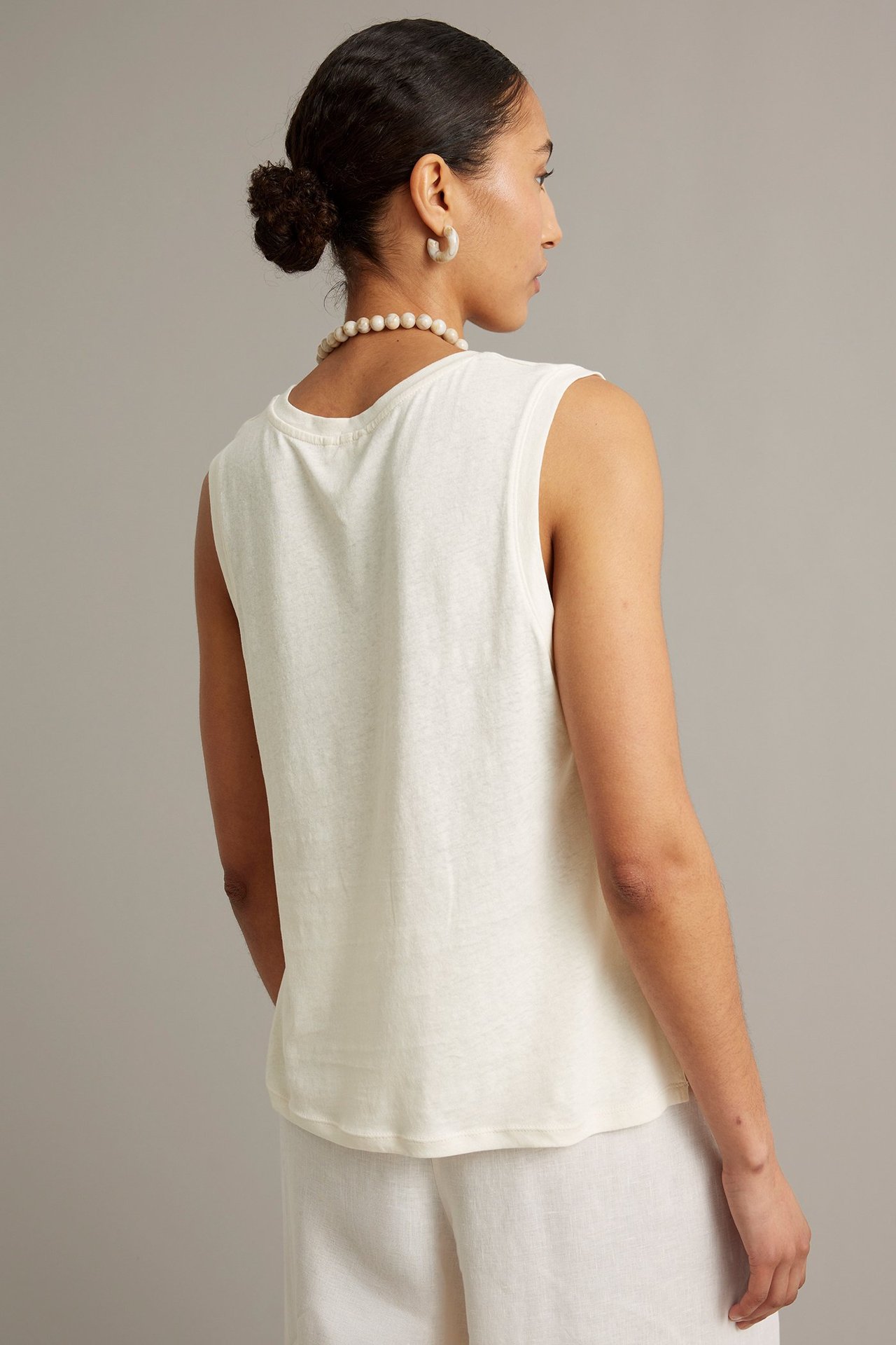 Bakåtvänd kvinna i vit linneblandad tanktopp, vita byxor, pärlhalsband och ringörhängen.