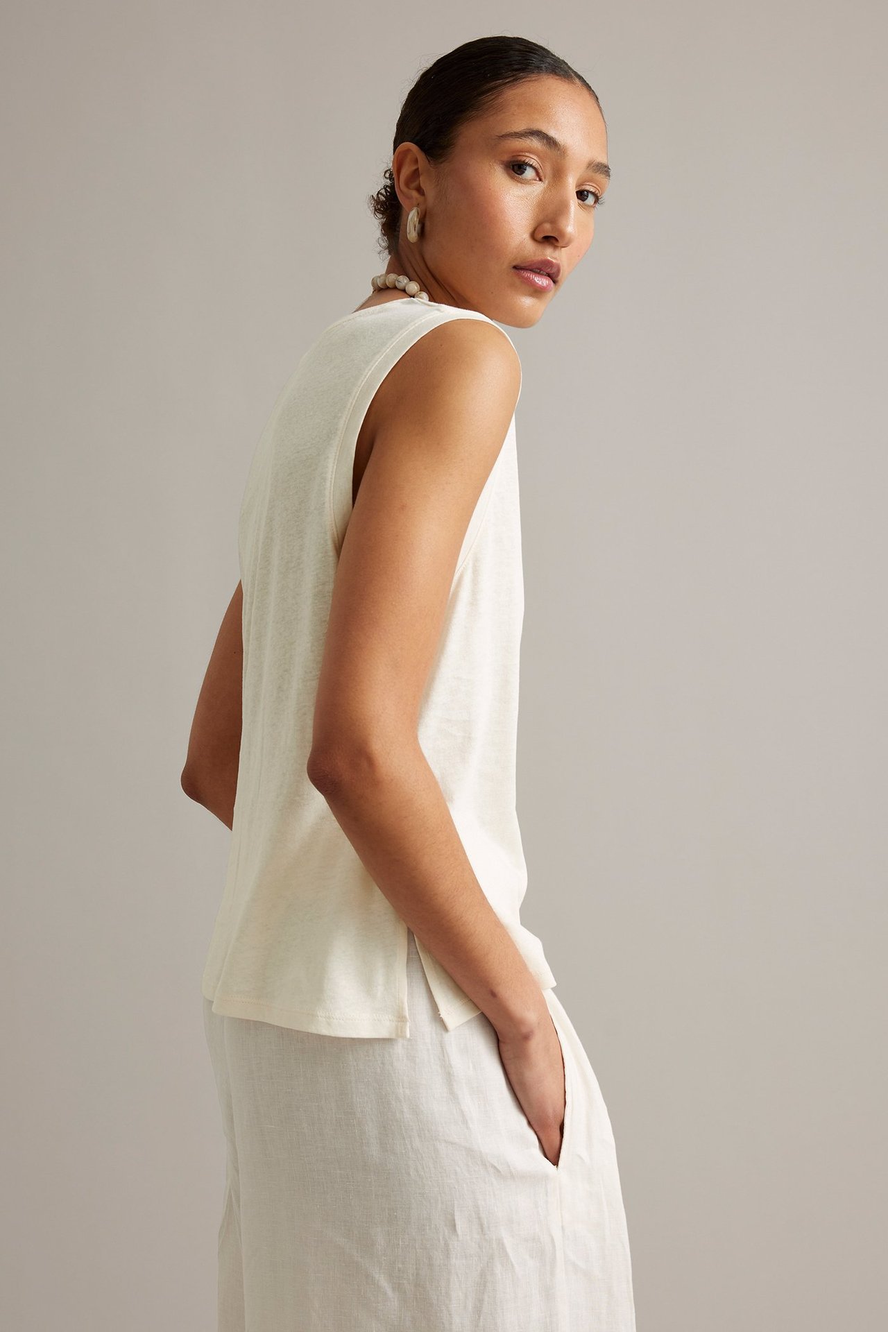 Sida-bakifrån vy av kvinna i vit ärmlös topp i linne/bomullsmix med slitsar och matchande byxor.