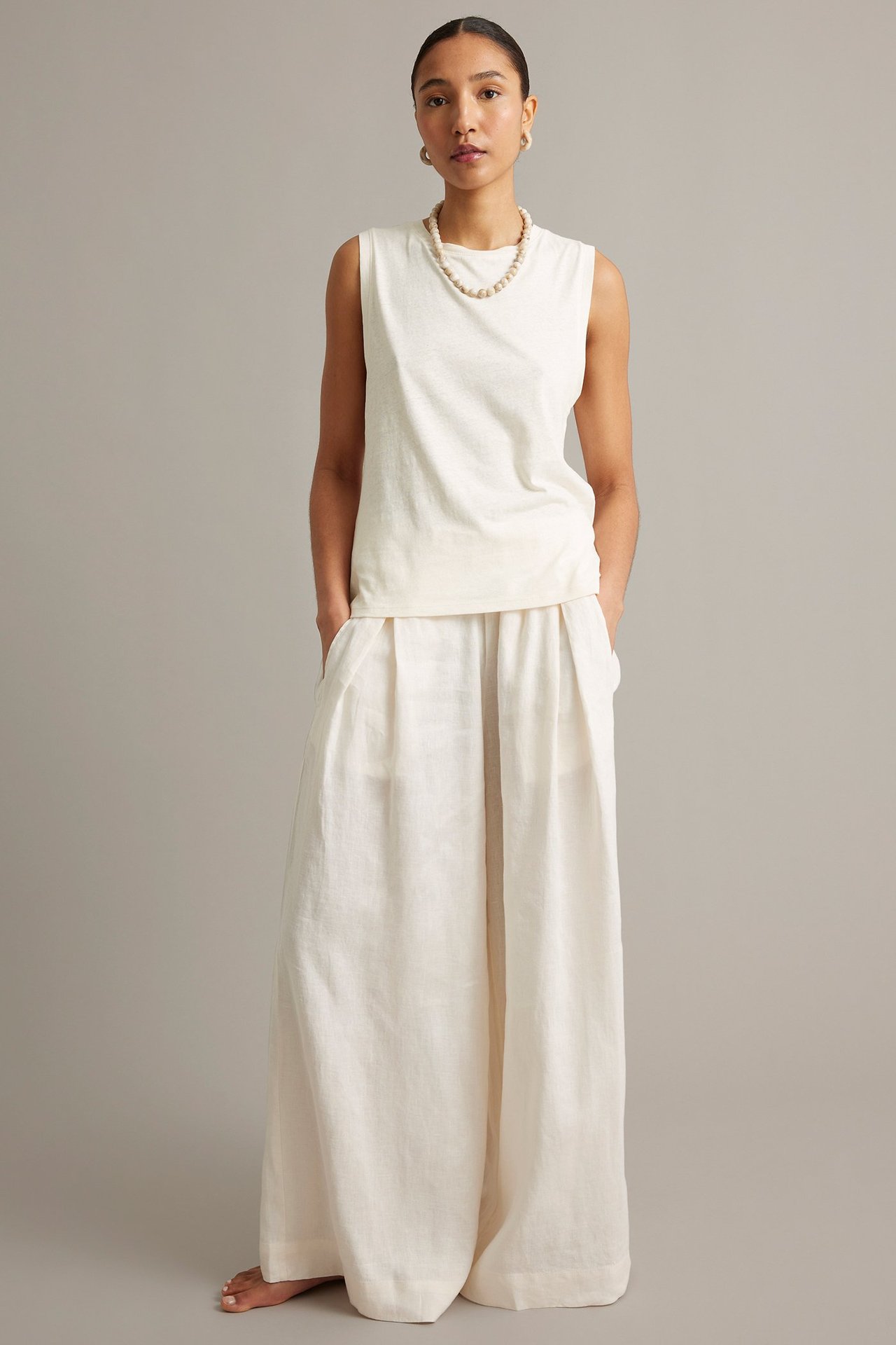 Framifrån, kvinna i vit ärmlös tanktopp i linneblandning, vida byxor, pärlhalsband och ringörhängen.