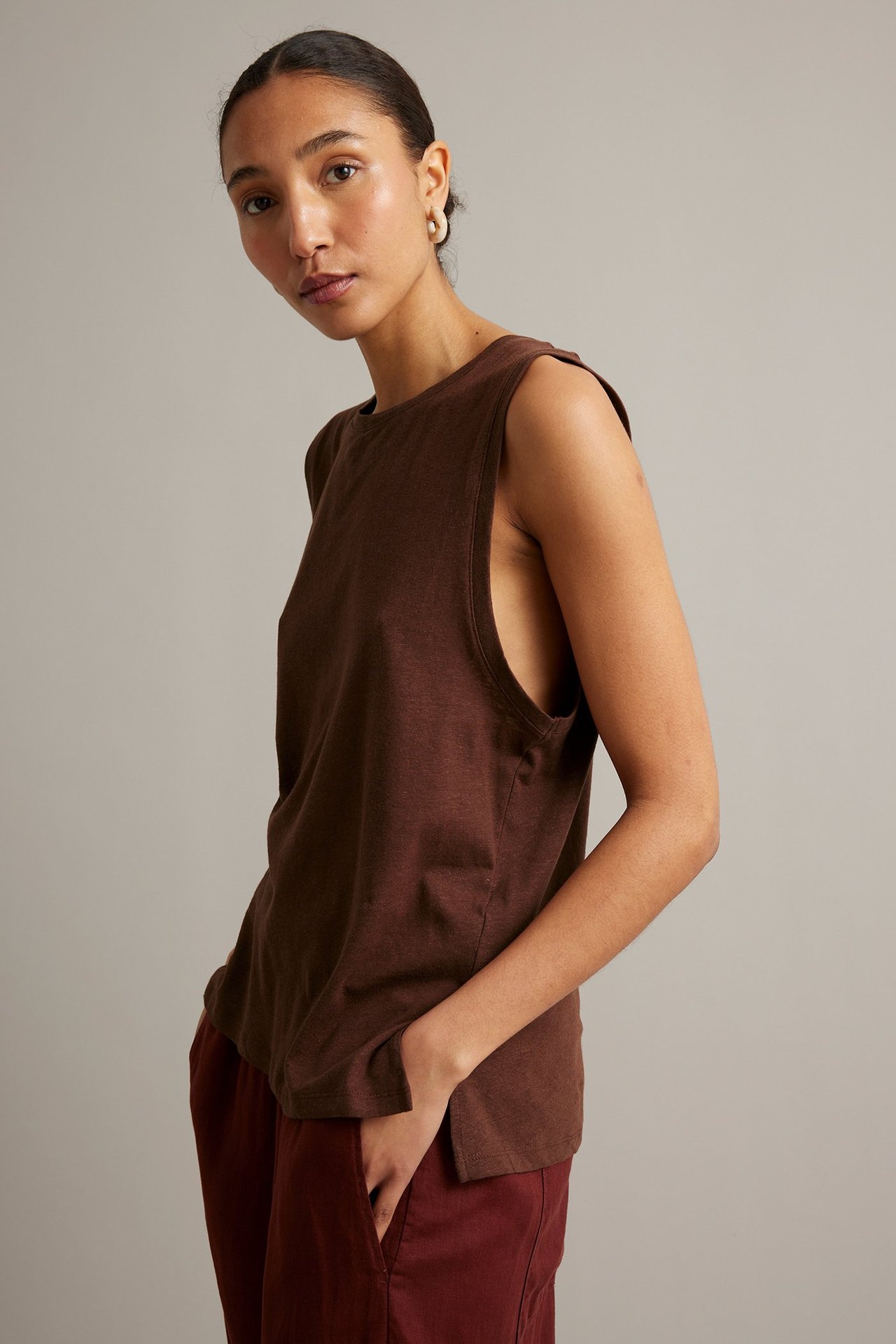 Frontvy: Kvinna i mörkbrun ärmlös topp i linne/bomull, mörkröda byxor och ljusa ringörhängen.