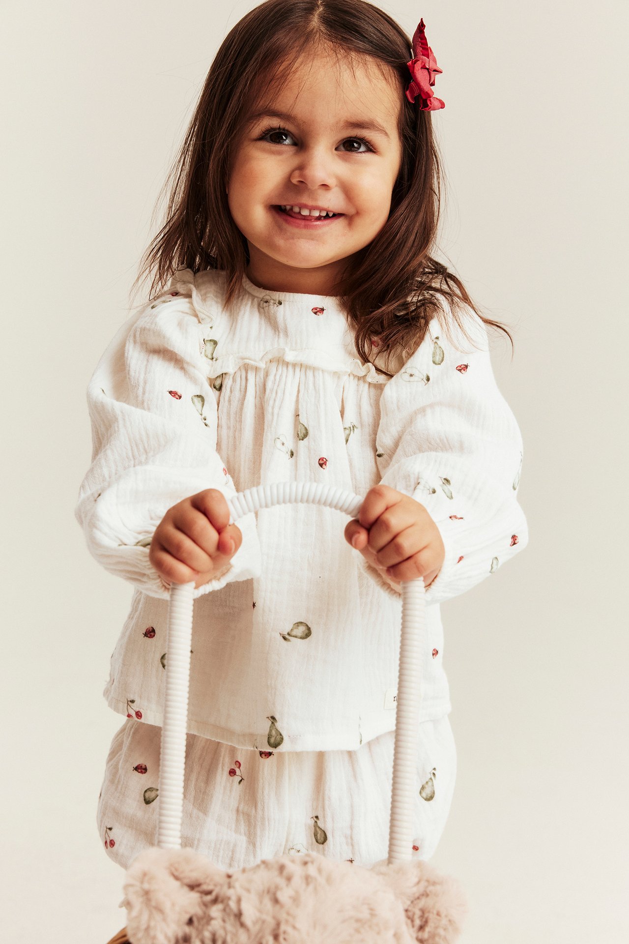 Edestä: Lapsi offwhite kuviollisessa musliinipuserossa ja yhteensopivissa shortseissa, pitelee pehmolelua.