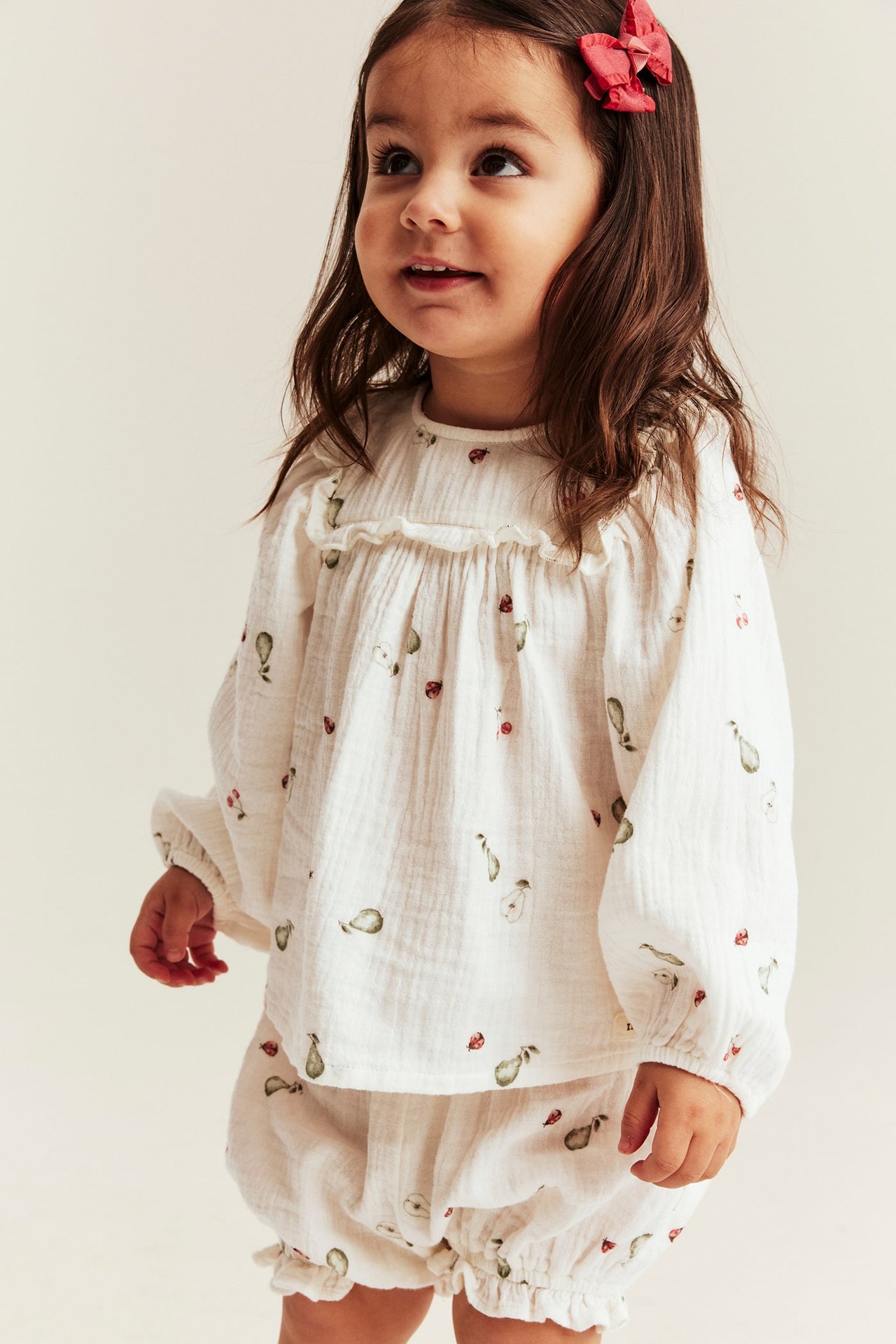 Edestäpäin: Pieni tyttö luonnonvalkoisessa kuviollisessa musliinisetissä, röyhelöpusero ja shortsit. Punainen rusetti.