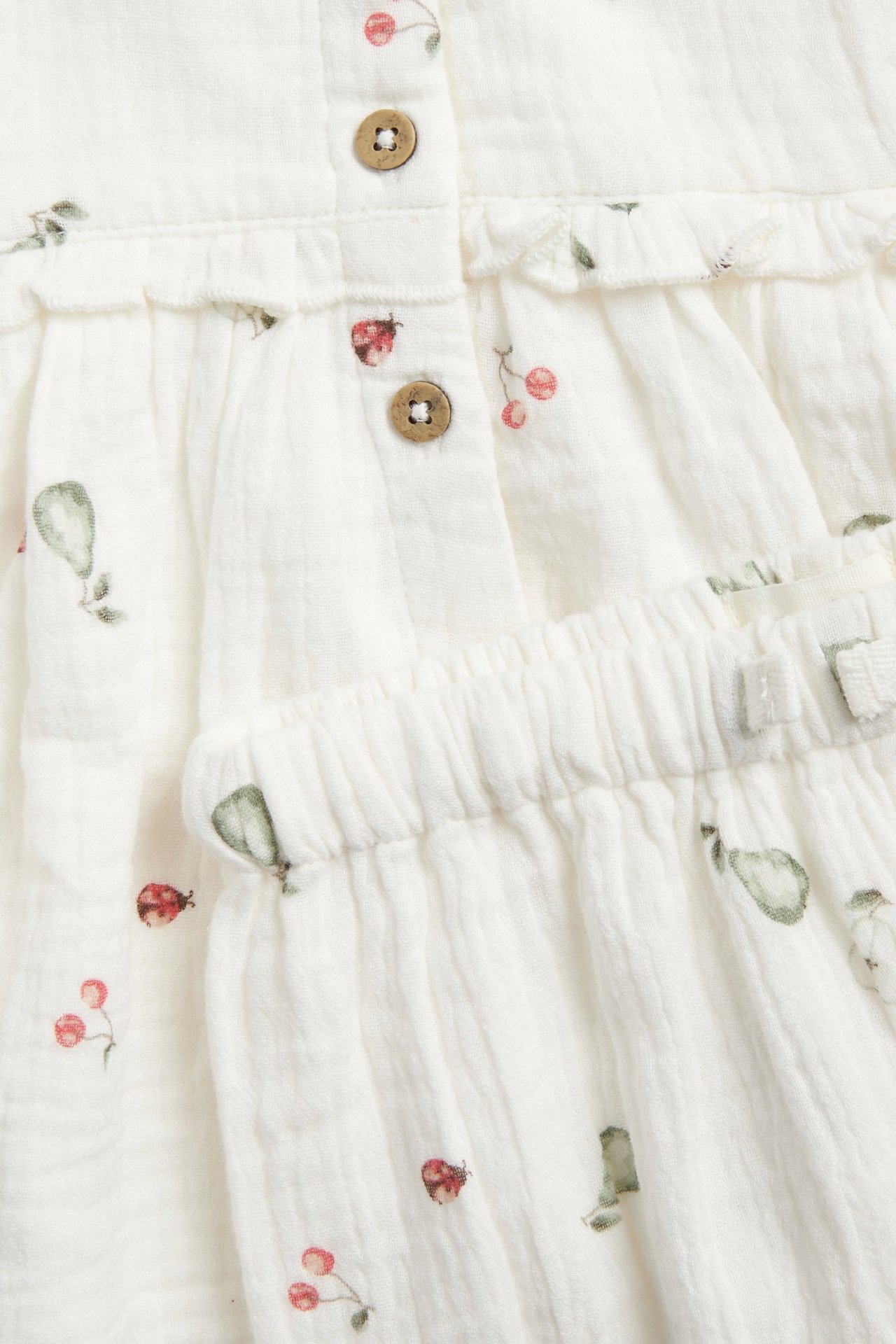 Zbliżenie na ecru muślinowy komplet z nadrukiem gruszek, wiśni i biedronek, z guzikami i falbankami.