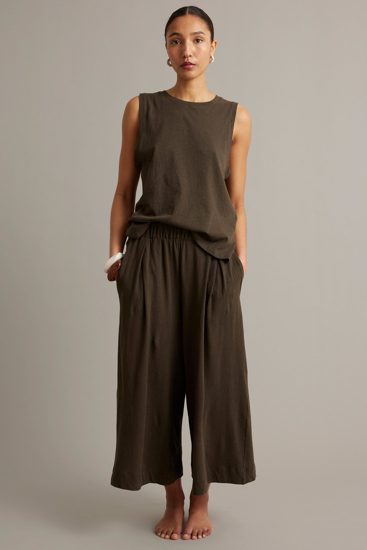 Framifrån, kvinna i bruna vida culottebyxor med veck, matchande ärmlös topp och ett vitt armband.
