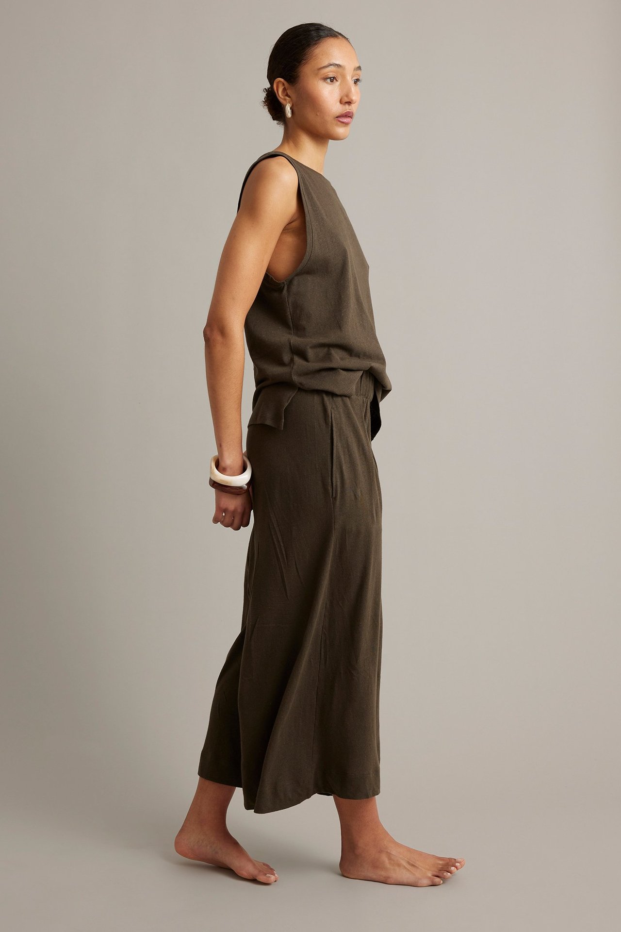 Sidovy av kvinna i mörkbruna vida culottebyxor med matchande ärmlös topp, armband och örhängen.