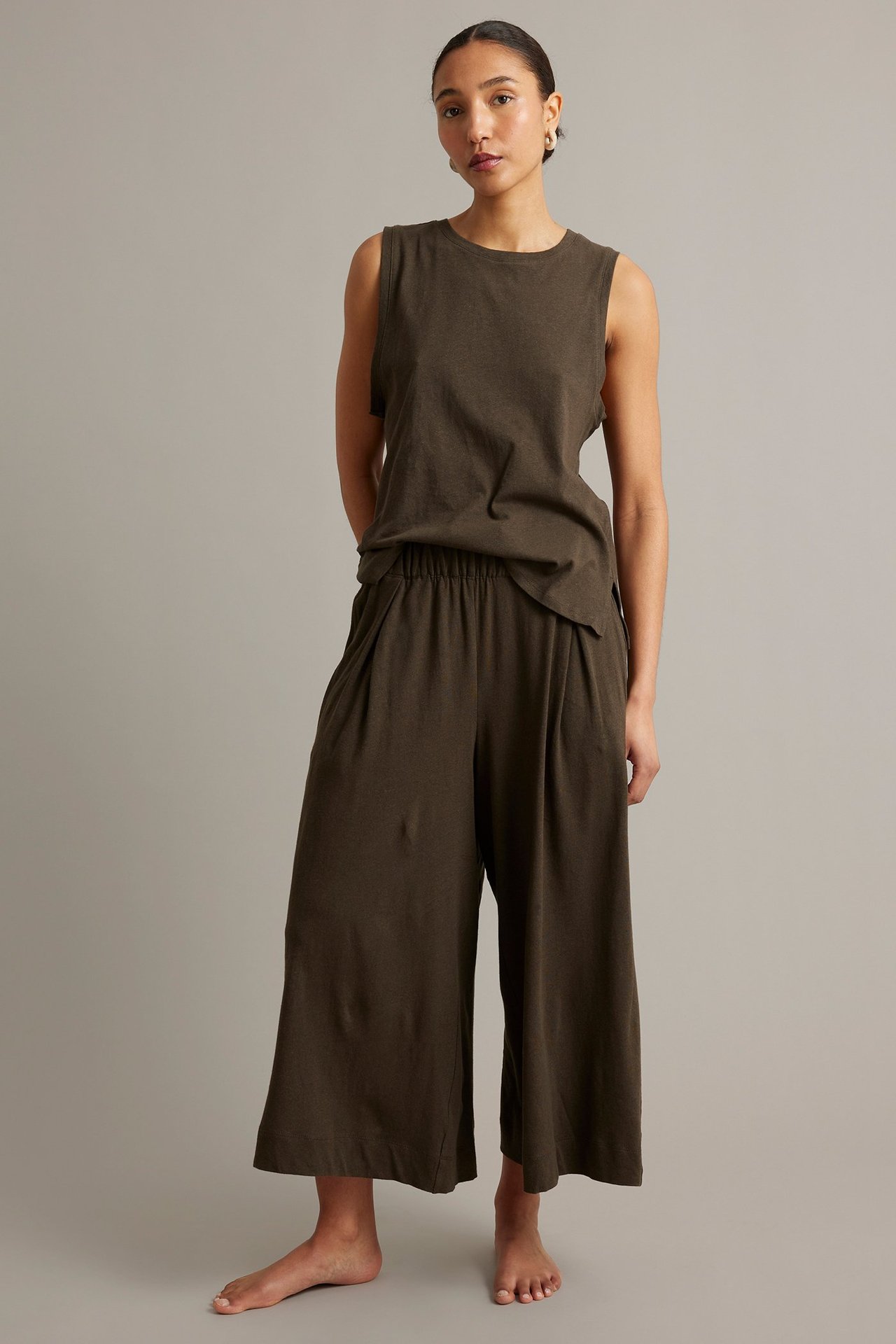 Widok z przodu: kobieta w brązowych, szerokich spodniach kulotach z zakładkami i pasującym topie bez rękawów.