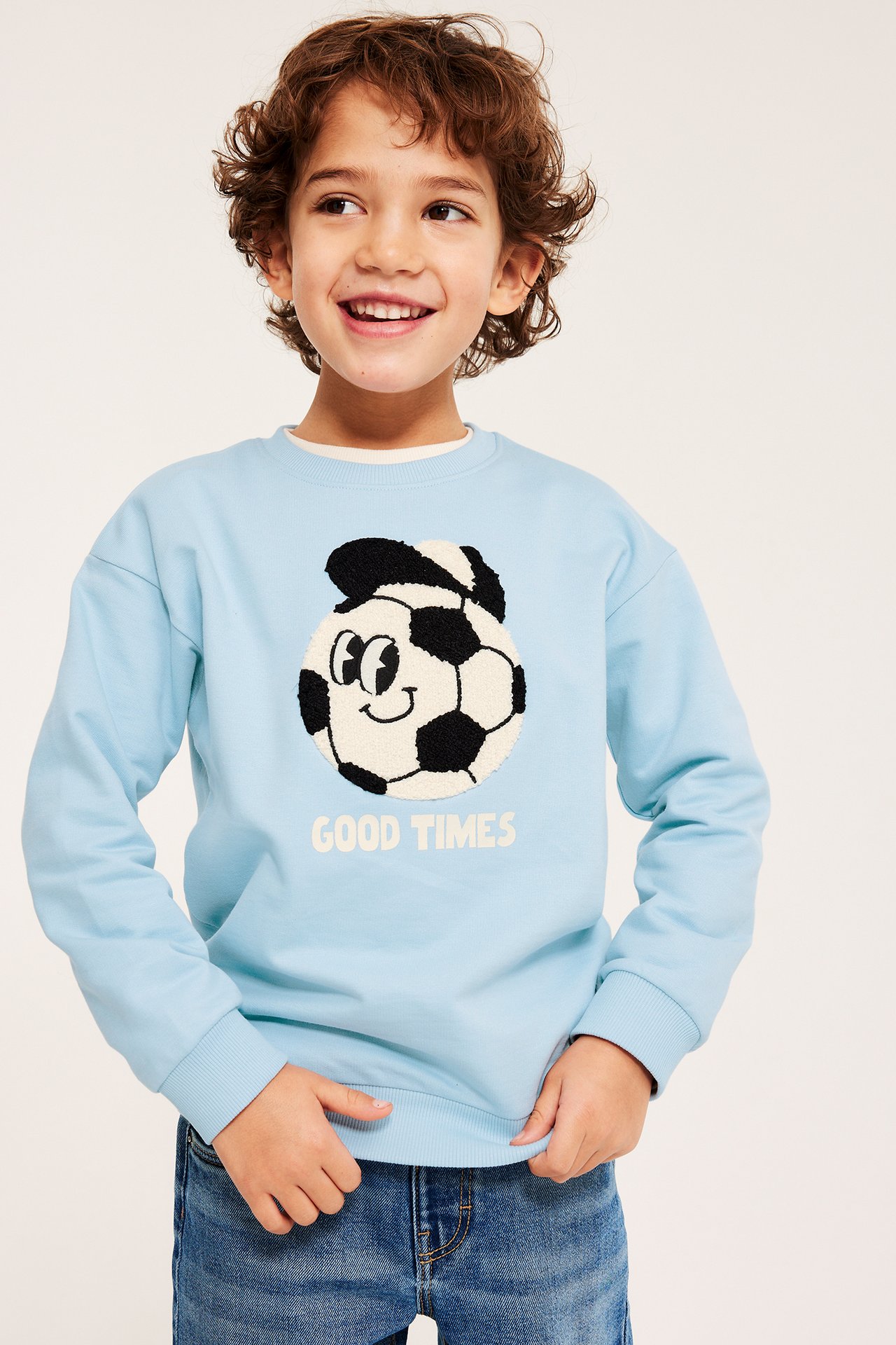 Edestä kuvattu lapsi yllään vaaleansininen puuvillainen collegepaita jalkapalloapplikaatiolla, sinisten farkkujen kanssa.