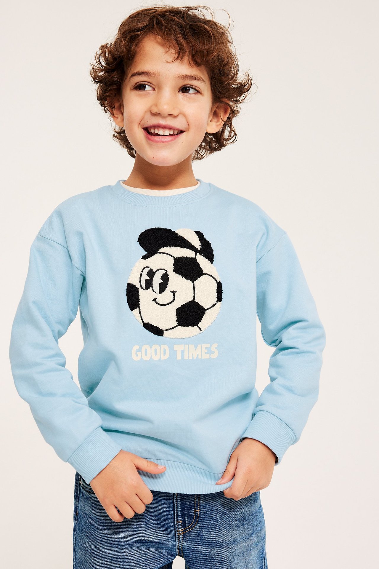 Edestä kuvattu lapsi yllään vaaleansininen puuvillainen collegepaita jalkapalloapplikaatiolla, sinisten farkkujen kanssa.