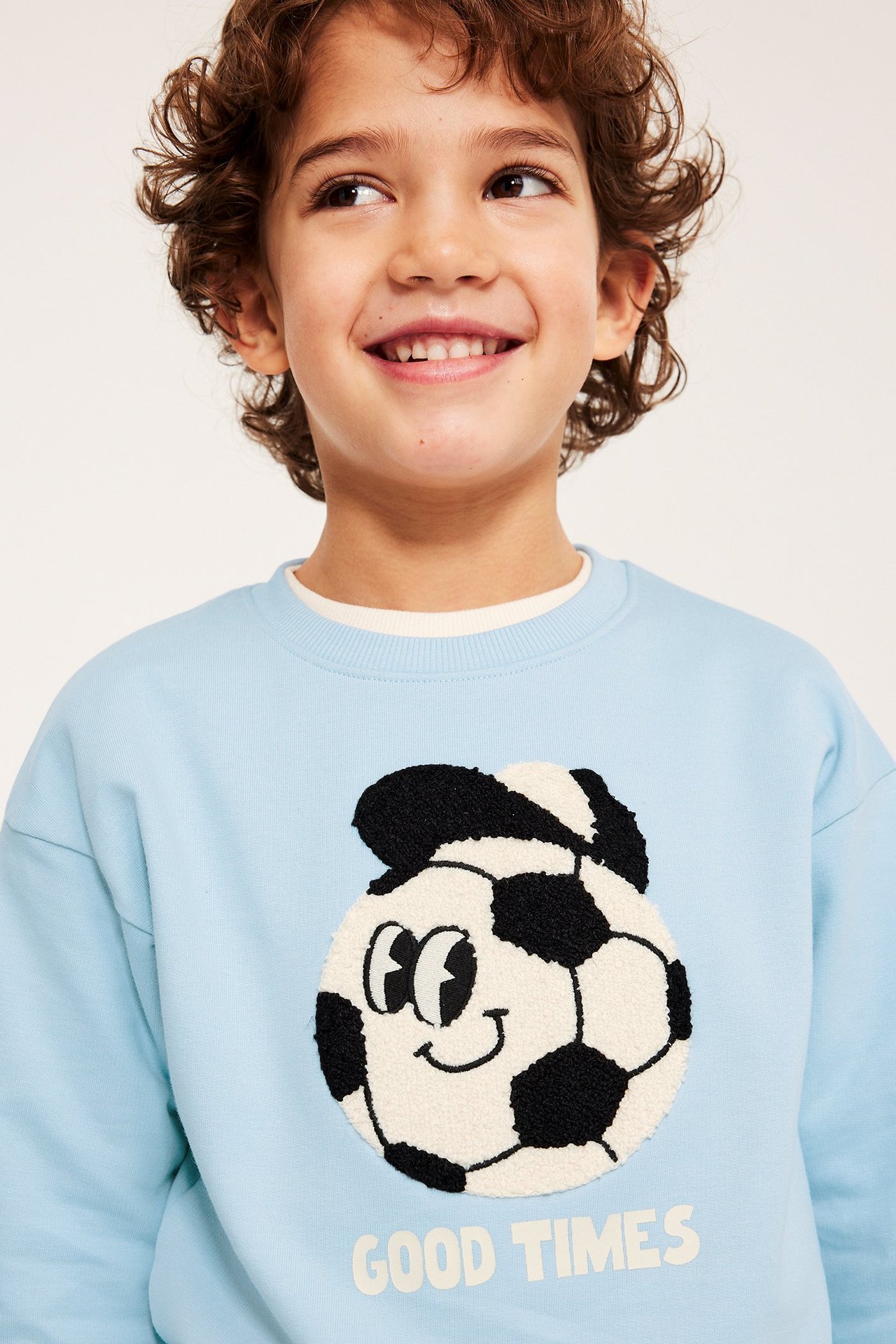Z przodu, uśmiechnięte dziecko w jasnoniebieskiej bluzie z aplikacją piłki i napisem "GOOD TIMES".