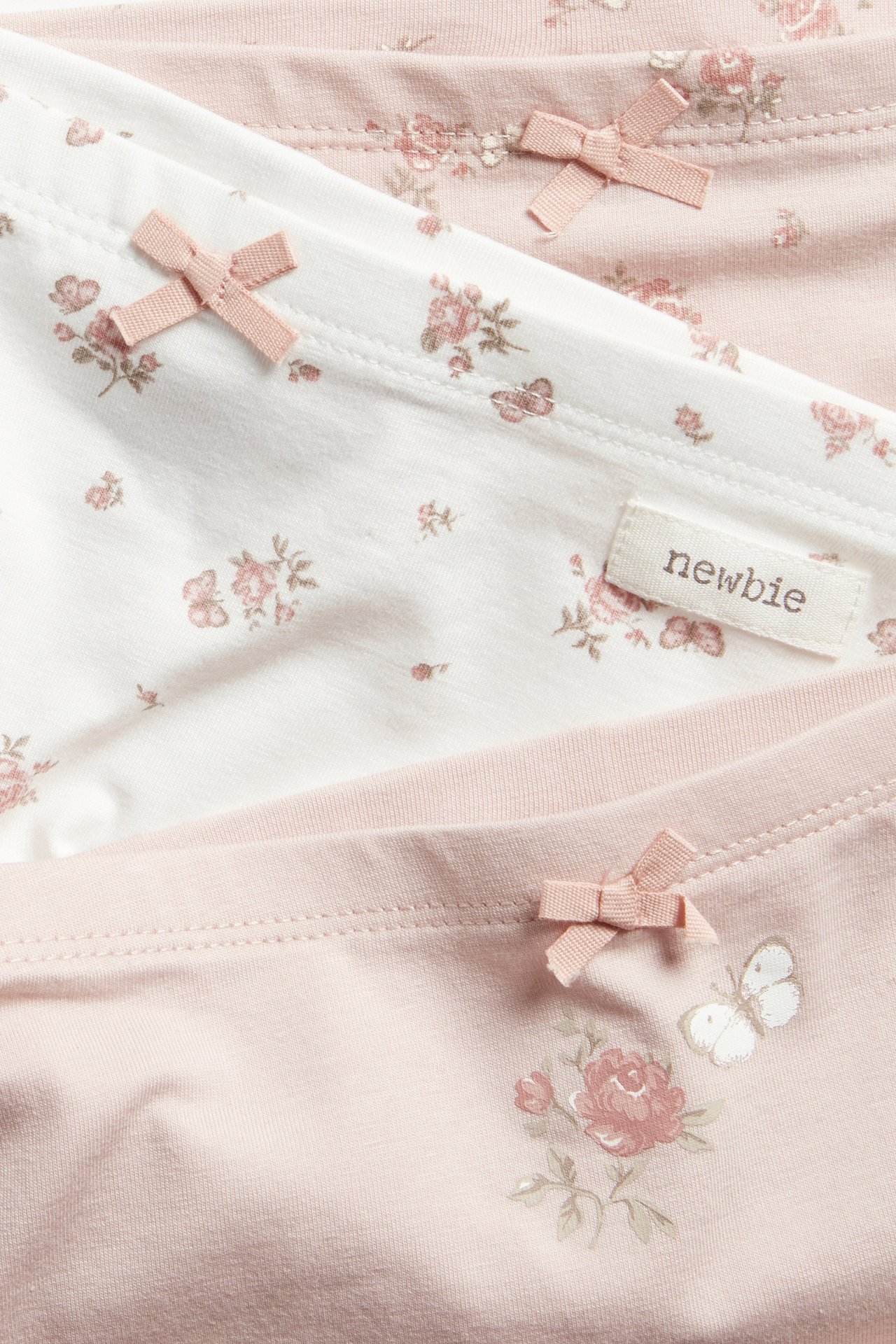 Zbliżenie na trzy dziecięce bokserki: białe i dwa różowe, w kwiatowy wzór, z małymi różowymi kokardkami.