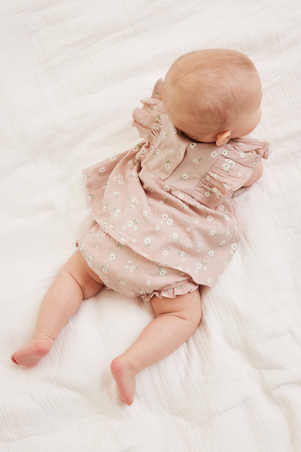 Vauva takaa: vaaleanpunainen kukkakuvioinen kreppisetti, jossa röyhelötoppi ja bloomerit.