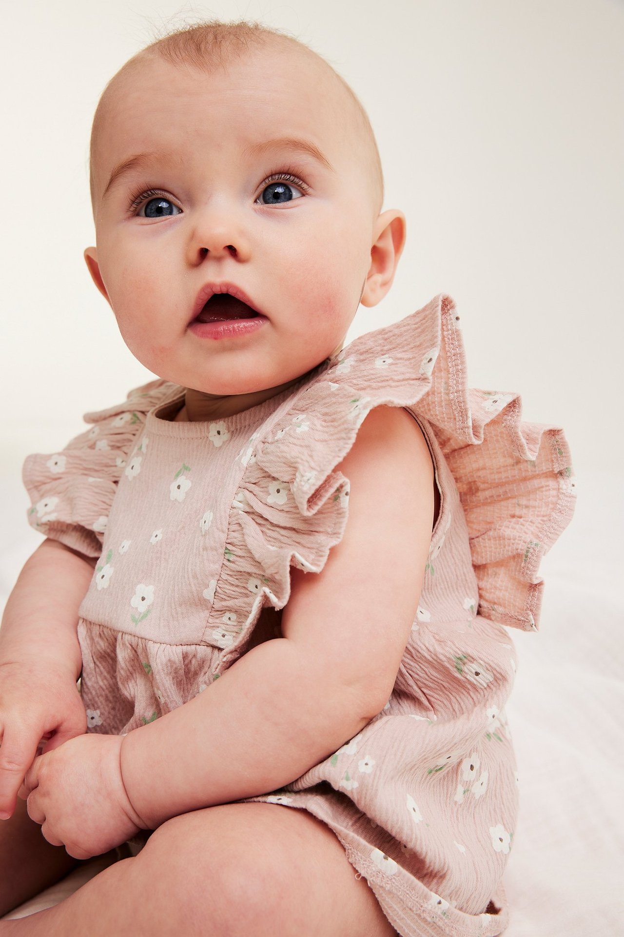 Lähikuva vauvasta, yllään vaaleanpunainen puuvillacrepesetti kukkakuviolla ja röyhelöolkapäillä.