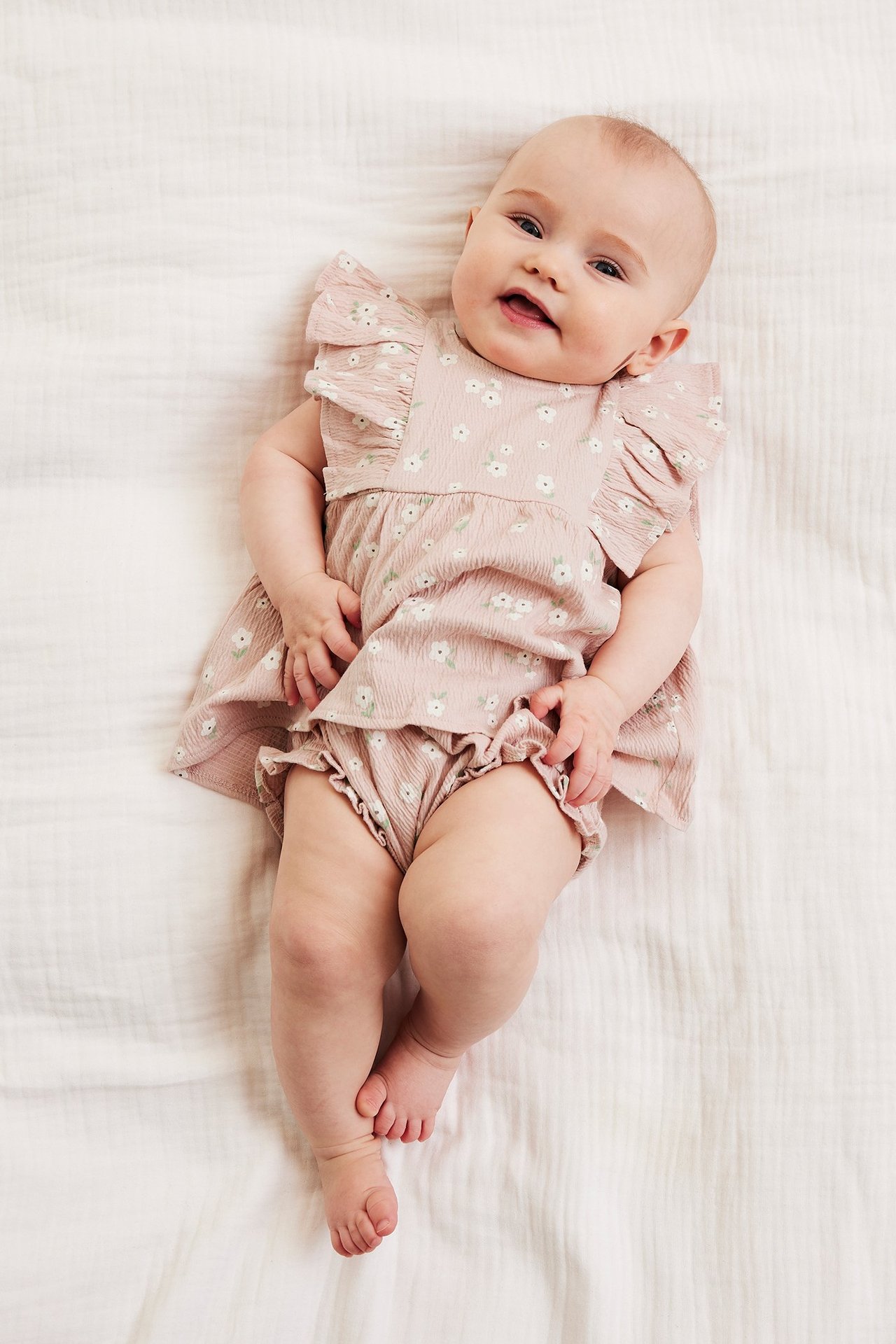 Yläkuva vauvasta, yllään vaaleanpunainen kukkakuvioinen kreppitoppi ja röyhelöbloomersit.