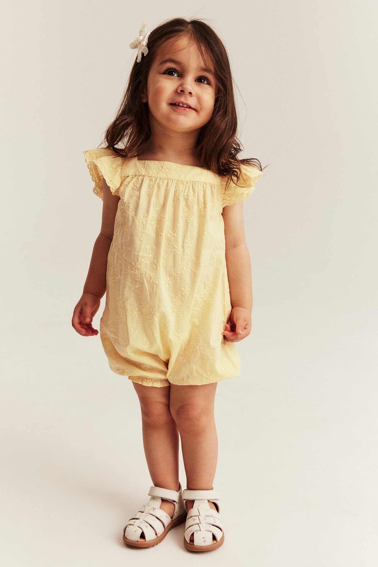 Edestäpäin: pikkutyttö yllään keltainen kirjailtu puuvillahaalari röyhelöhihoilla ja vaaleabeiget sandaalit.