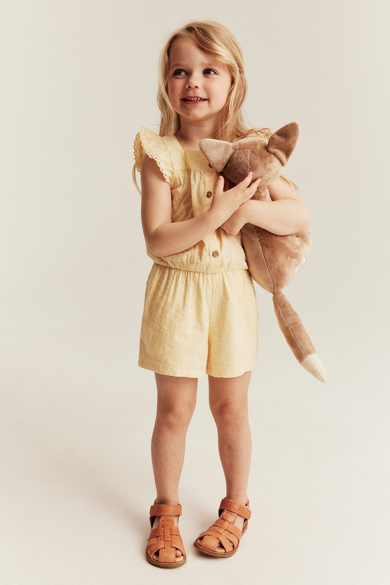 Edestä: Lapsi keltaisessa kirjaillussa haalarissa, röyhelöhihat, ruskea pehmolelu ja ruskeat sandaalit.