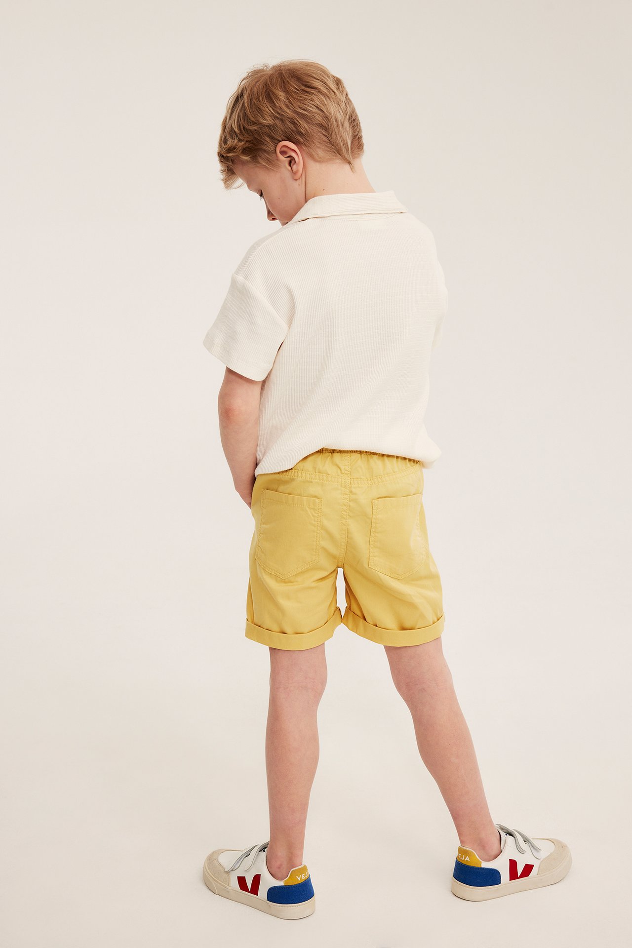 Widok z tyłu: dziecko w żółtych bawełnianych szortach z elastyczną talią, kremowym polo i biało-czerwono-niebieskich sneakersach.