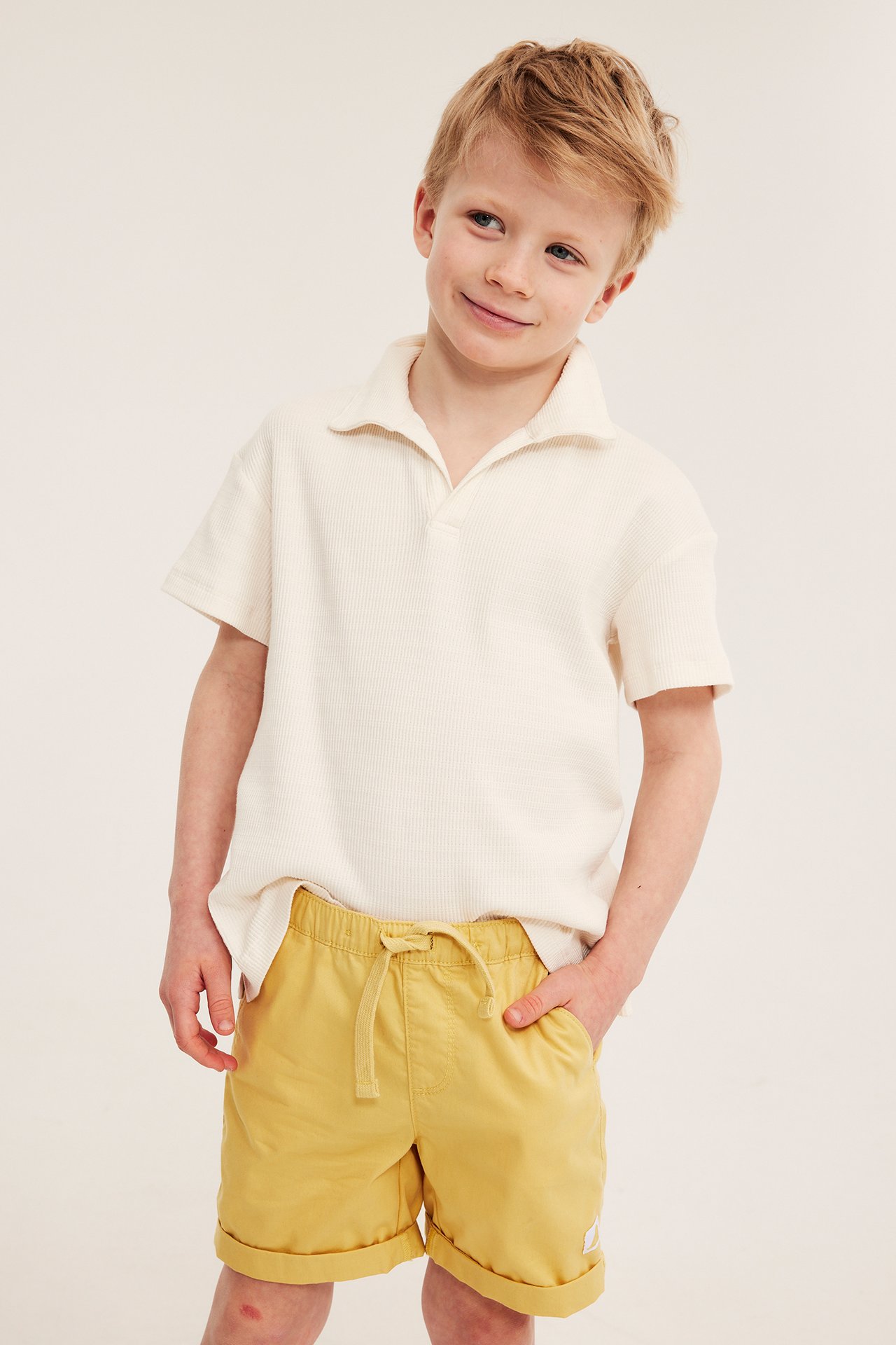 Edestä: Lapsi keltaisissa puuvillatvillishortseissa, joissa kiristysnyöri, ja luonnonvalkoisessa pikeepaidassa.