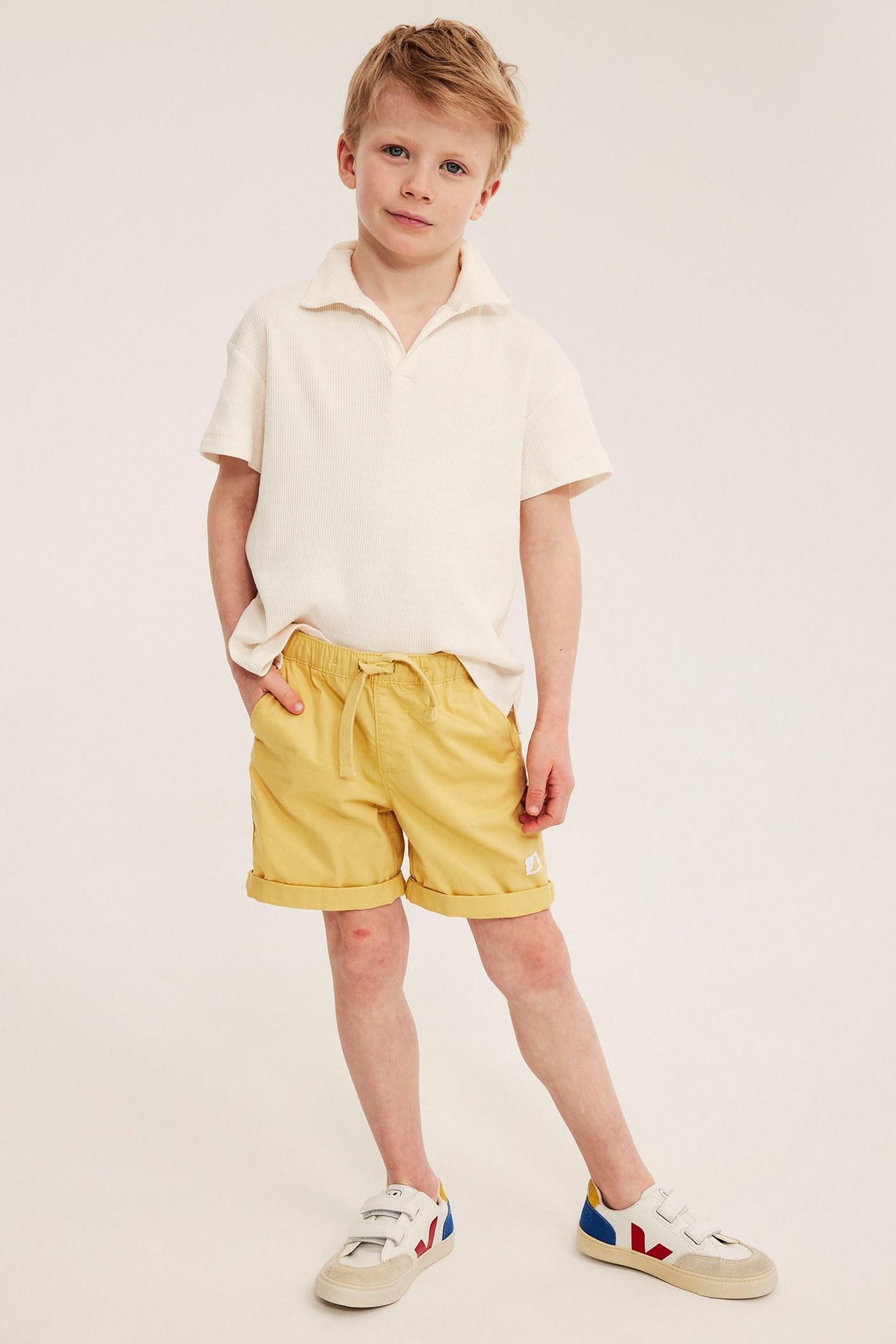 Widok z przodu dziecka w żółtych bawełnianych szortach, kremowym t-shircie polo i białych sneakersach.