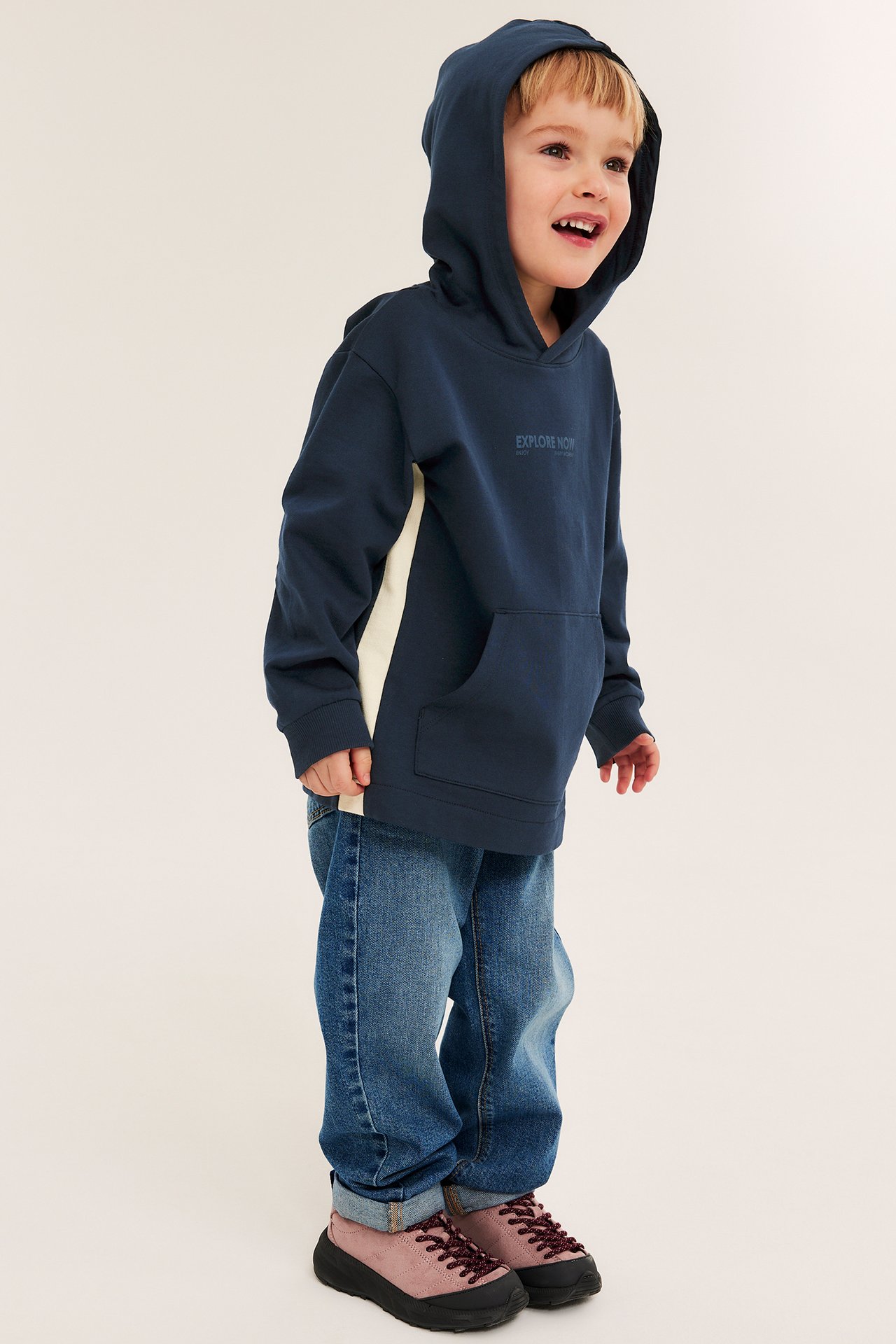 Sidebilde av et barn i mørkeblå bomullshettegenser med kremfarget sidepanel, blå jeans og rosa snøresko.