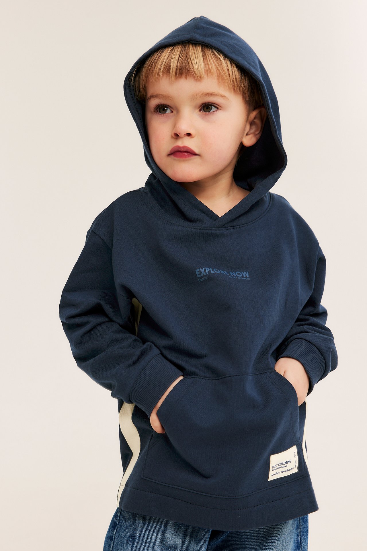 Widok z przodu: Dziecko w granatowej bawełnianej bluzie z kapturem, tekstem, kontrastowymi panelami bocznymi i kieszenią kangurką, w niebieskich dżinsach.