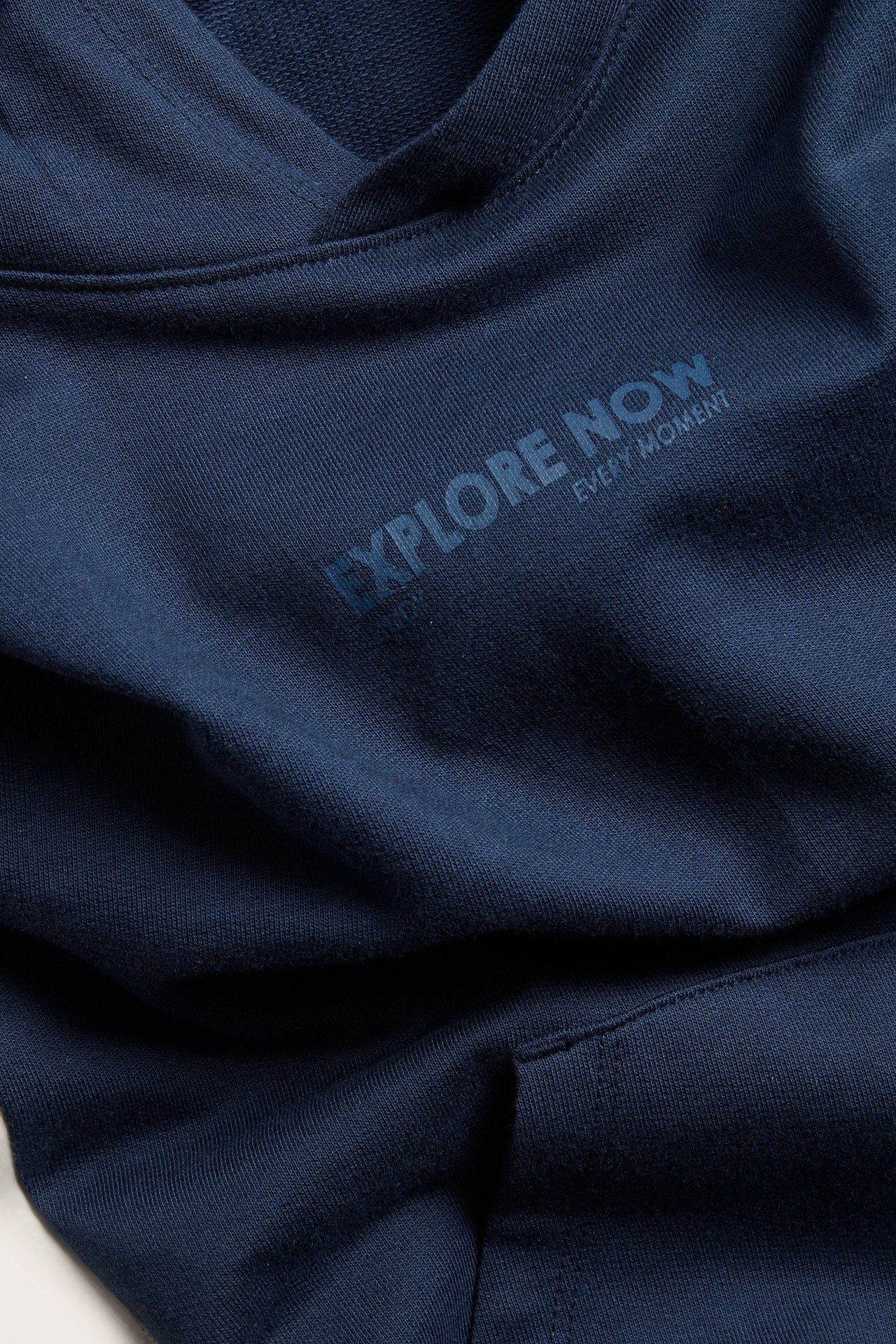 Lähikuva edestä lapsen tummansinisestä puuvillahupparista, jossa on kengurutasku ja 'EXPLORE NOW' -painatus.