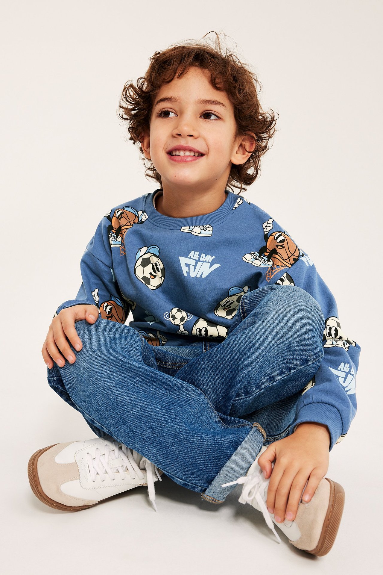 Edestä, lapsi sinisessä kuvioidussa collegepaidassa urheiluaiheilla, farkuissa ja valkoisissa tennareissa.