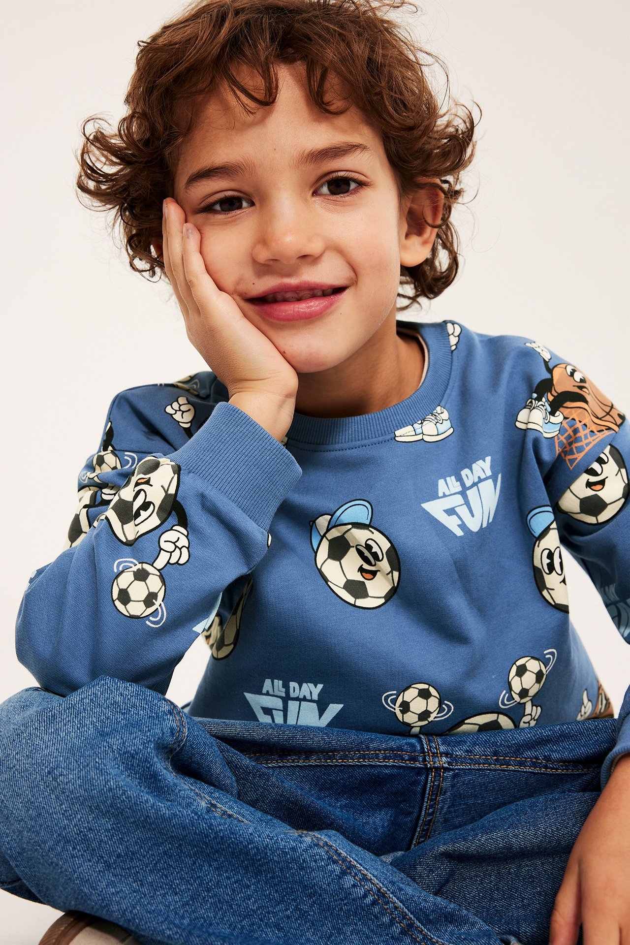 Närbild framifrån på ett barn som bär en blå sweatshirt i bomull med fotbollsmönster och blå jeans.