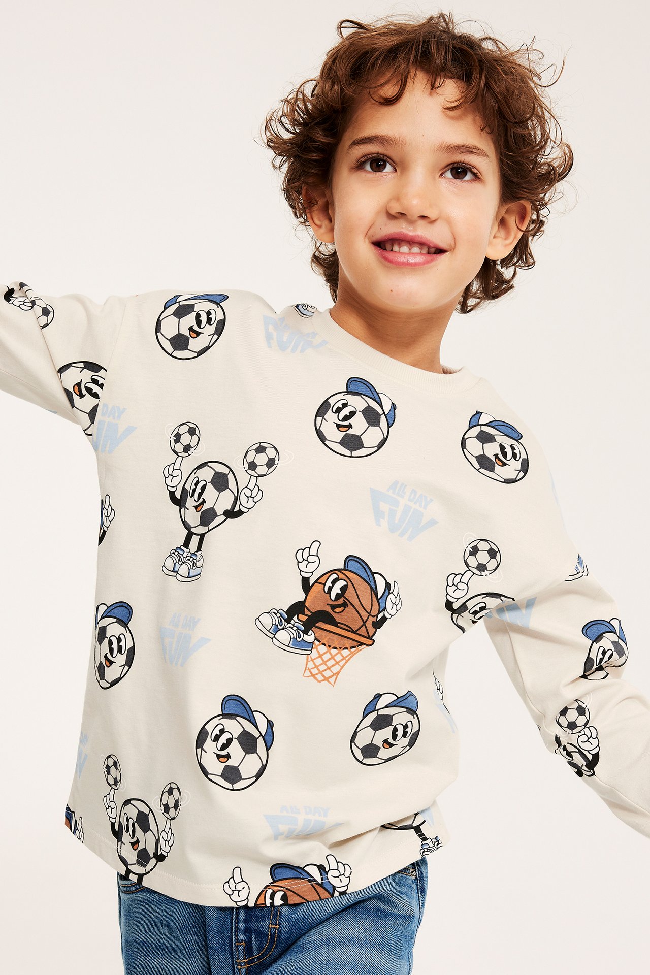 Widok z przodu: Dziecko w jasnobeżowej bawełnianej koszulce z długim rękawem z nadrukiem piłek sportowych i dżinsach.