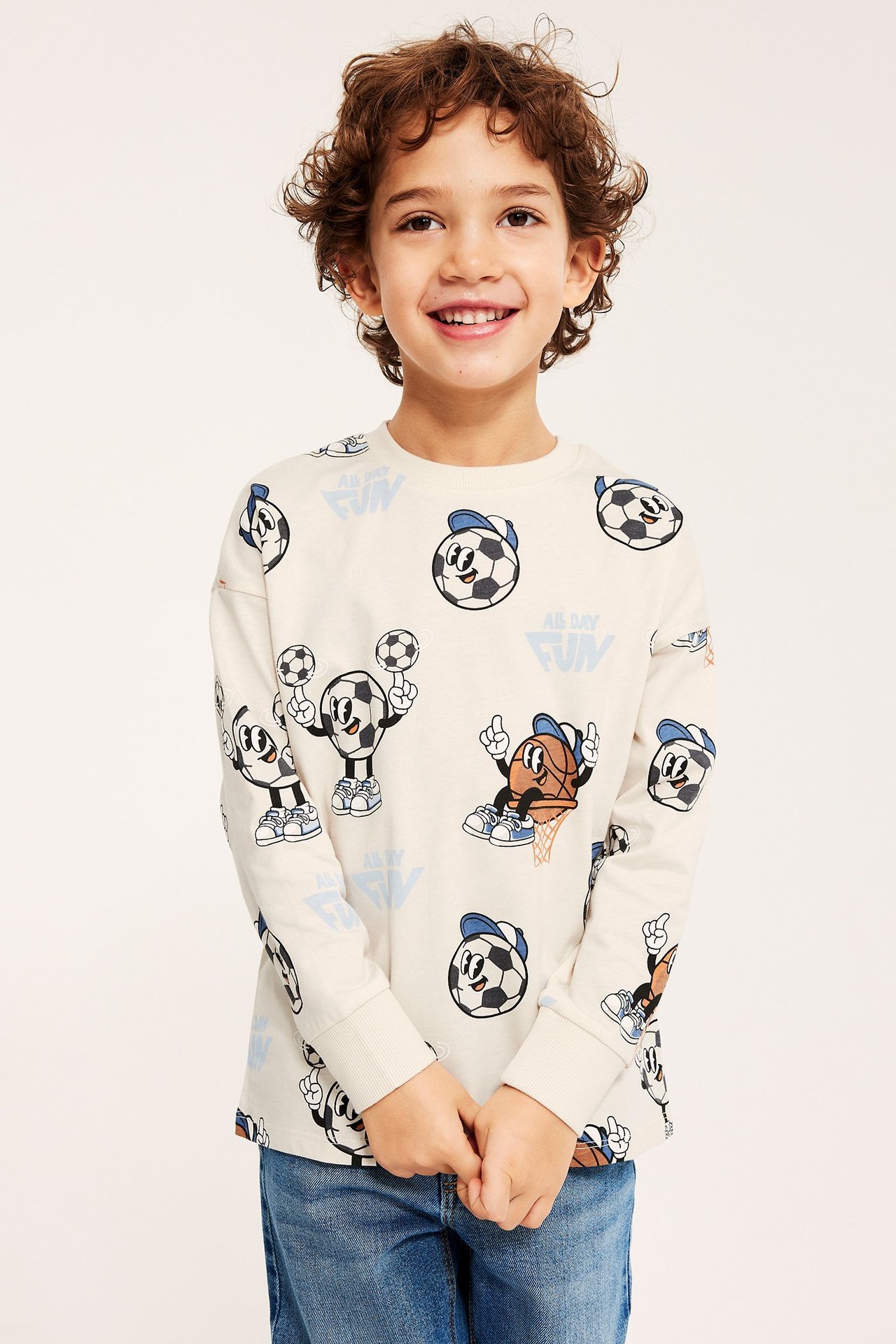 Przód uśmiechniętego dziecka w jasnobeżowej, długiej bawełnianej koszulce z nadrukiem piłek i niebieskich dżinsach.