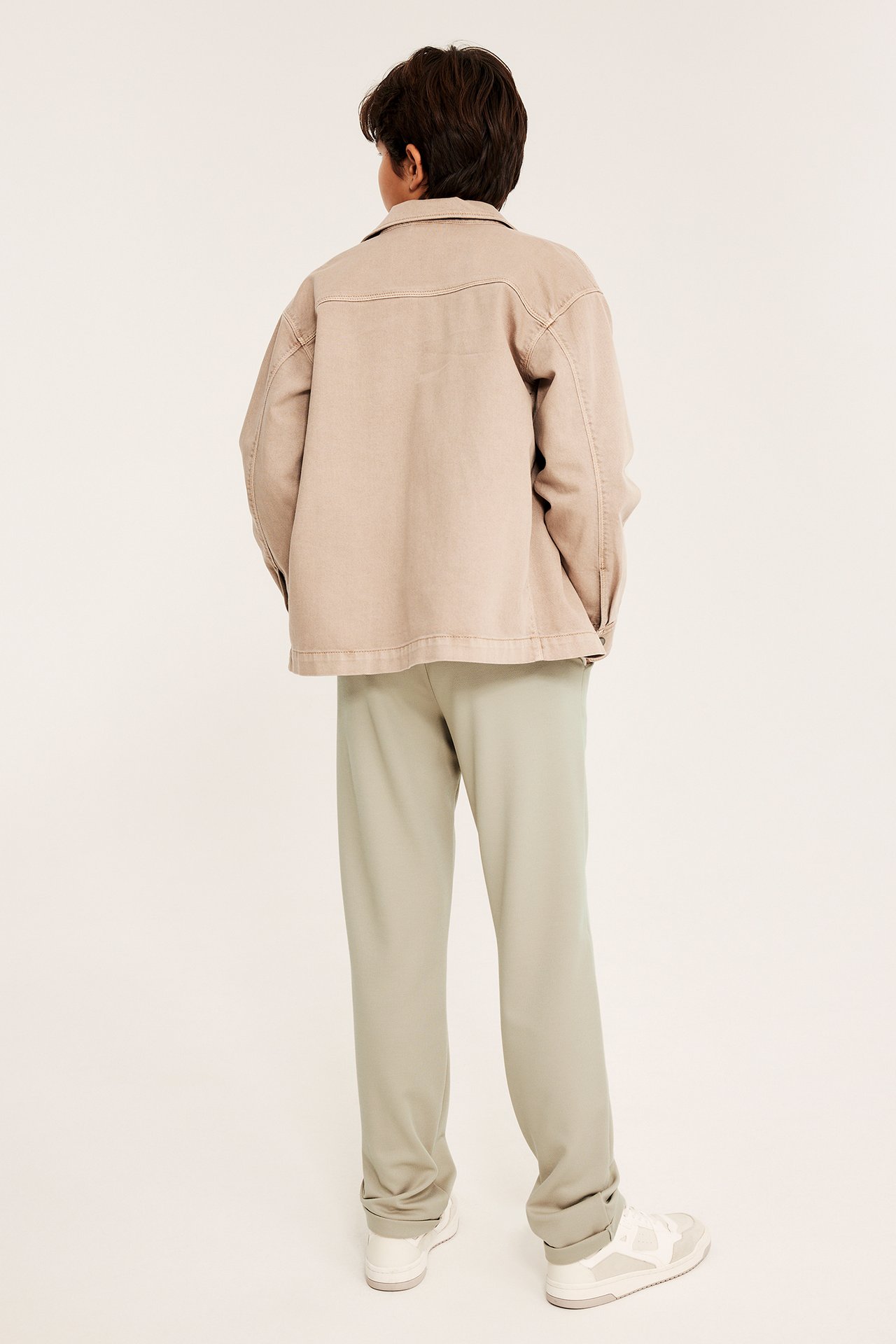 Bakfra, et barn i lysegrønne dressbukser, en beige oversized skjortejakke og hvite joggesko.