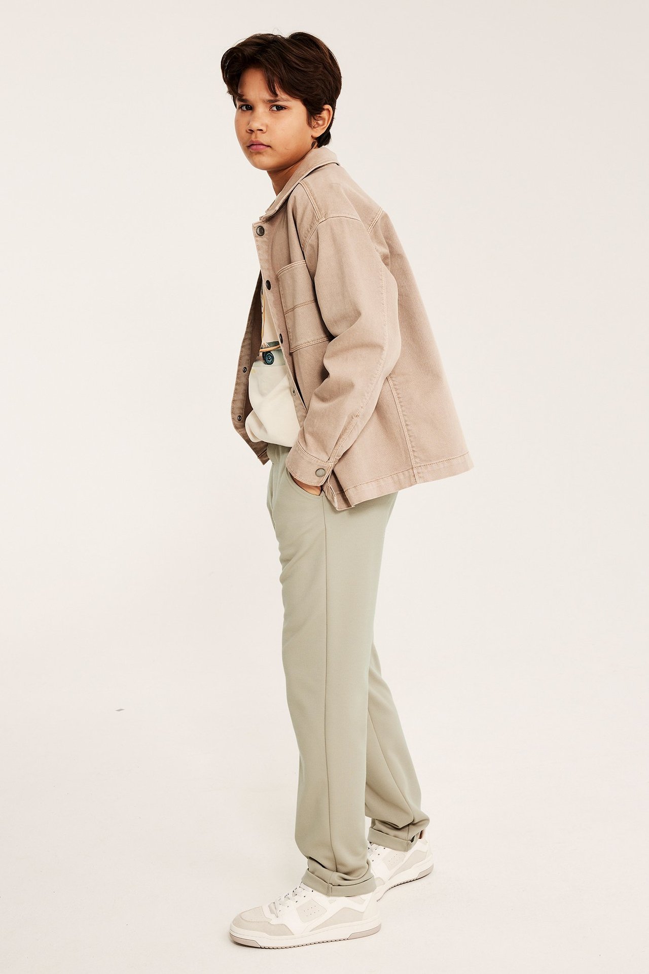 Sivukuva: Lapsi vaaleanvihreissä suorissa pukuhousuissa, beigessä ylipaidassa, valkoisessa t-paidassa ja valkoisissa lenkkareissa.