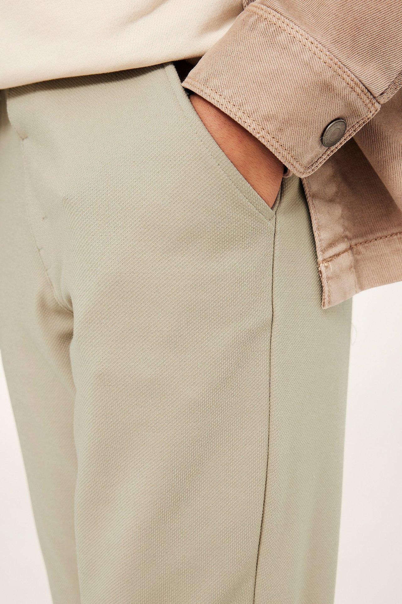 Nærbilde: Barns beige dressbukser med hånd i lommen, en lys topp og en lysebrun jakke.