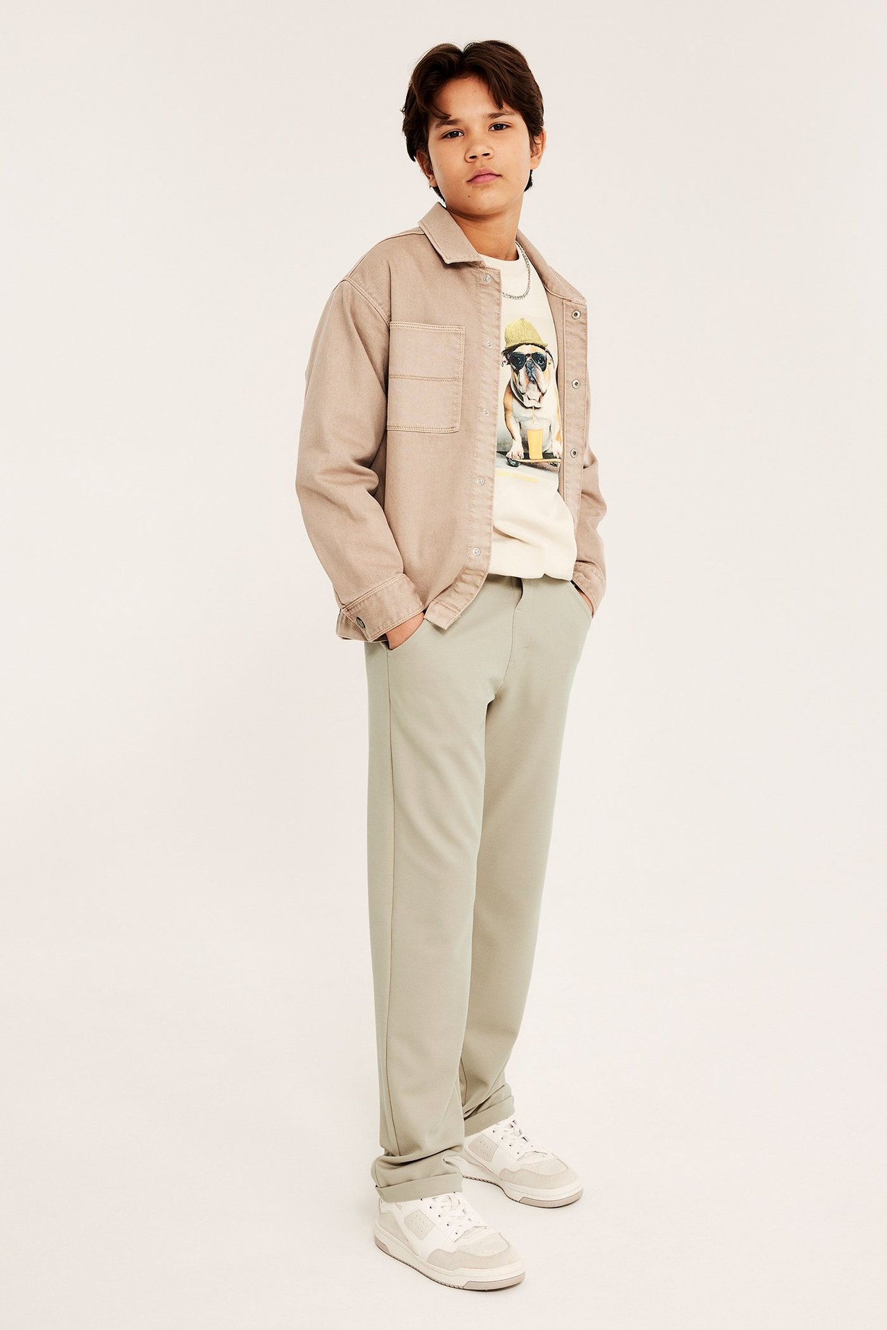 Widok z przodu: Dziecko w jasnozielonych spodniach wizytowych, otwartej beżowej koszuli, koszulce z grafiką i sneakersach.