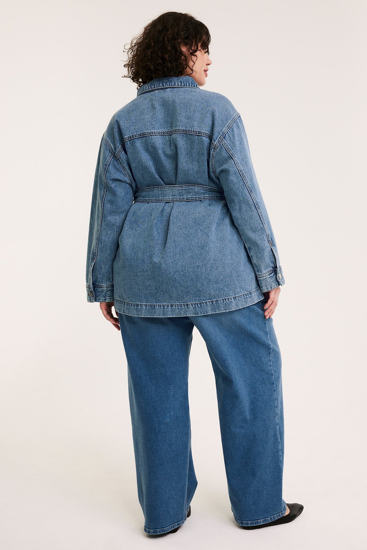 Naisen takakuva vaaleansinisessä farkkutakissa solmitulla vyöllä, sekä leveät farkkuhousut.