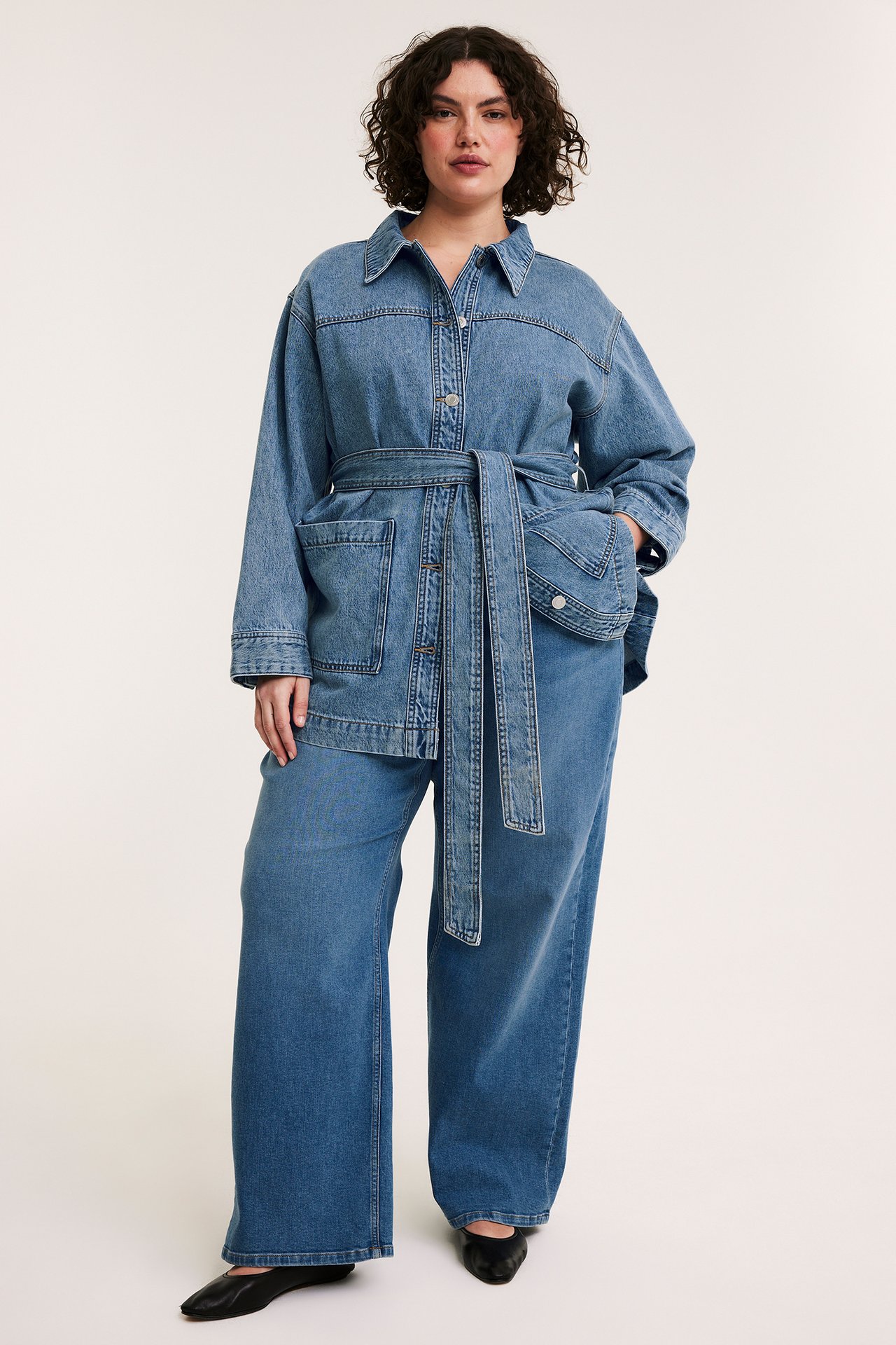 Kvinne i lysblå denim shacket med knyttet belte, matchende vide jeans og svarte flats, forfra.