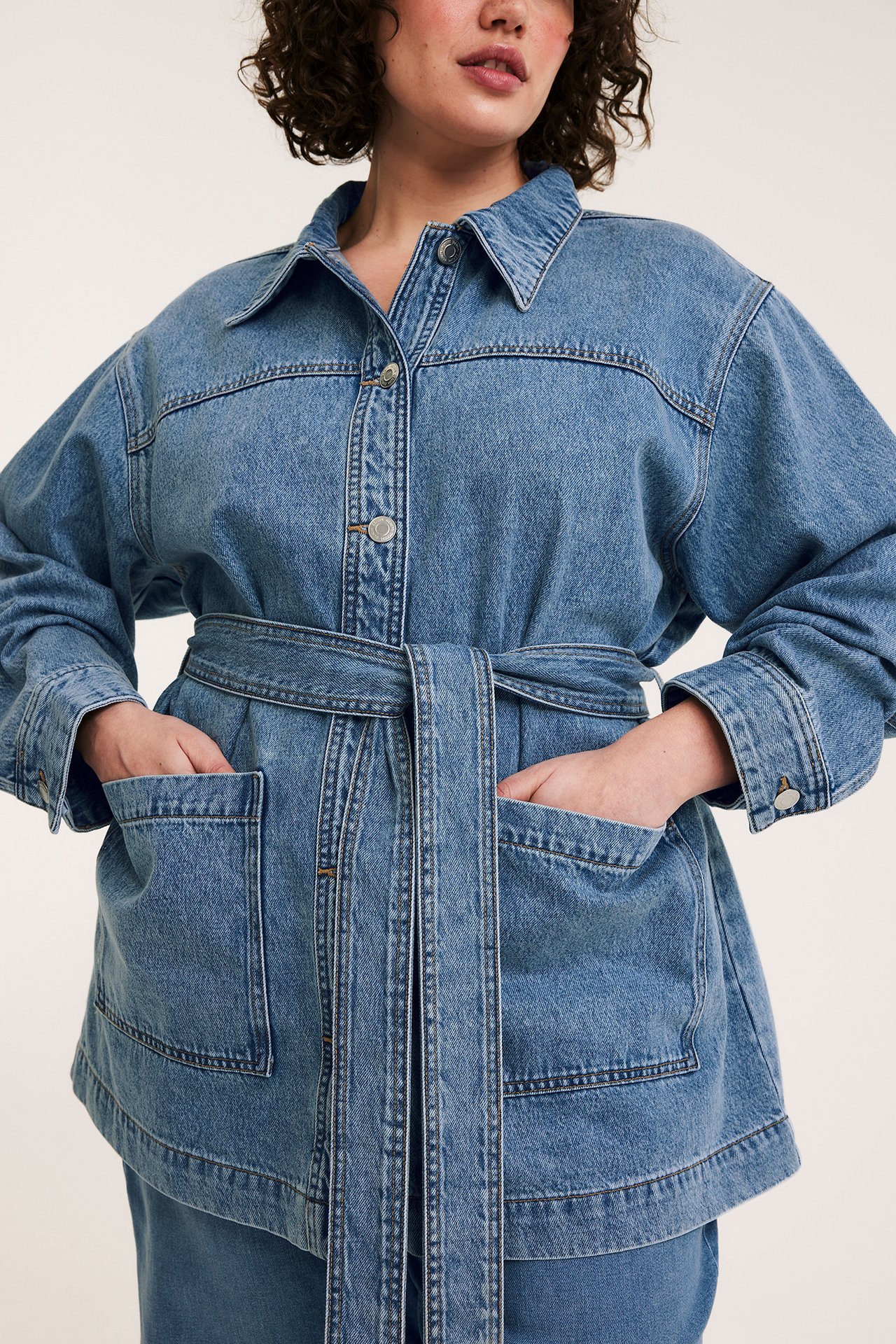 Naisen etunäkymä vaaleansinisessä denim-shaketissa, jossa napit, paikkataskut ja yhteensopiva solmittava vyö.